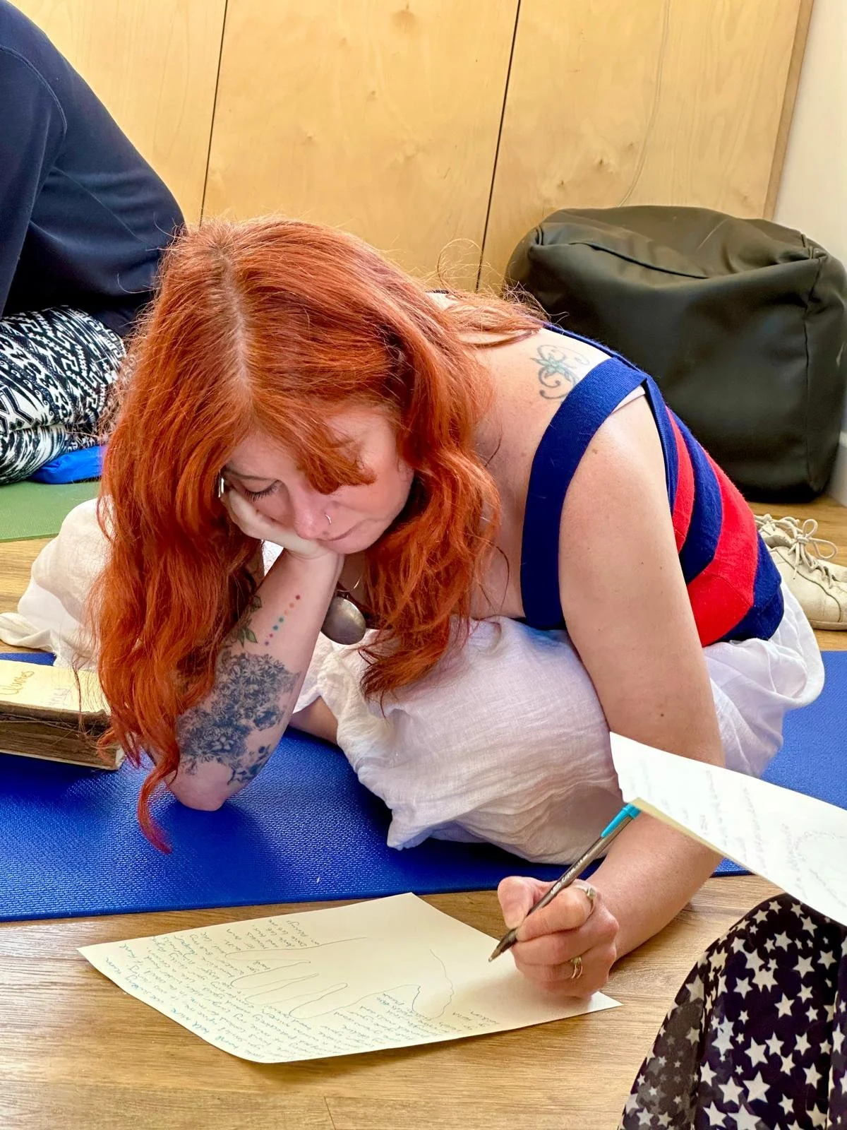 A woman with red hair and tattoos is lying face down on a yoga mat, writing on a large piece of paper. She appears to be in a relaxed mood, resting her head on her hand while writing with a pen. There are books and a backpack nearby in a cozy indoor setting.