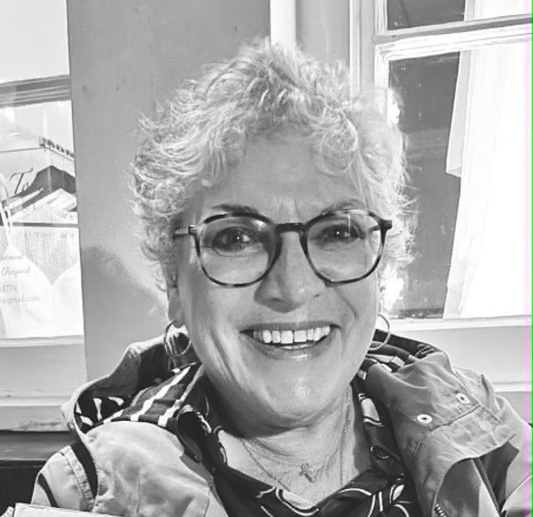 A smiling woman with short curly hair, glasses, and earrings, sitting near a window in a casual setting.