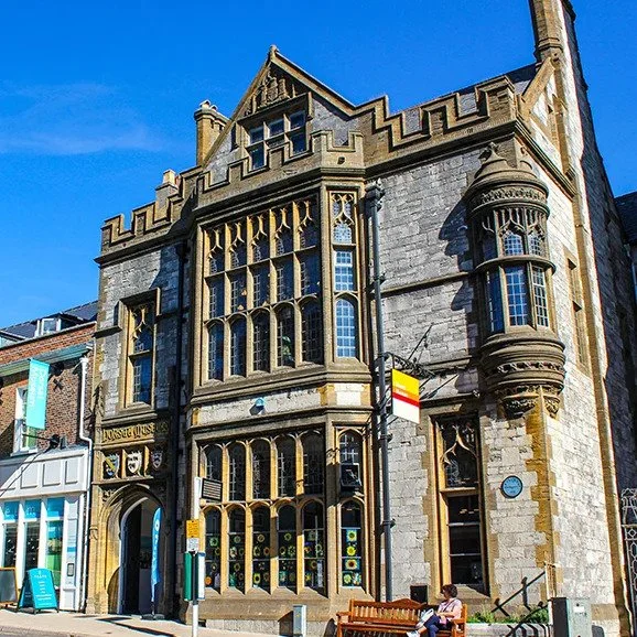 A historic stone building with large stained glass windows, ornate architectural details, and a turret, situated on a sunny street with people sitting on a bench nearby.
