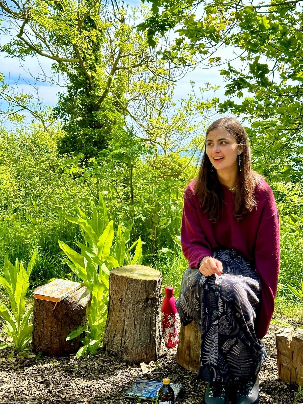 A young woman with long brown hair wearing a maroon sweater and patterned skirt sitting on a small wooden stool outdoors in a green, leafy forest clearing. She is smiling and looking to the side. There are tree stumps in front of her with a book and a bottle placed on top of them. A bottle of red water is beside her. Bright green plants and trees surround her, with a blue sky visible in the background.