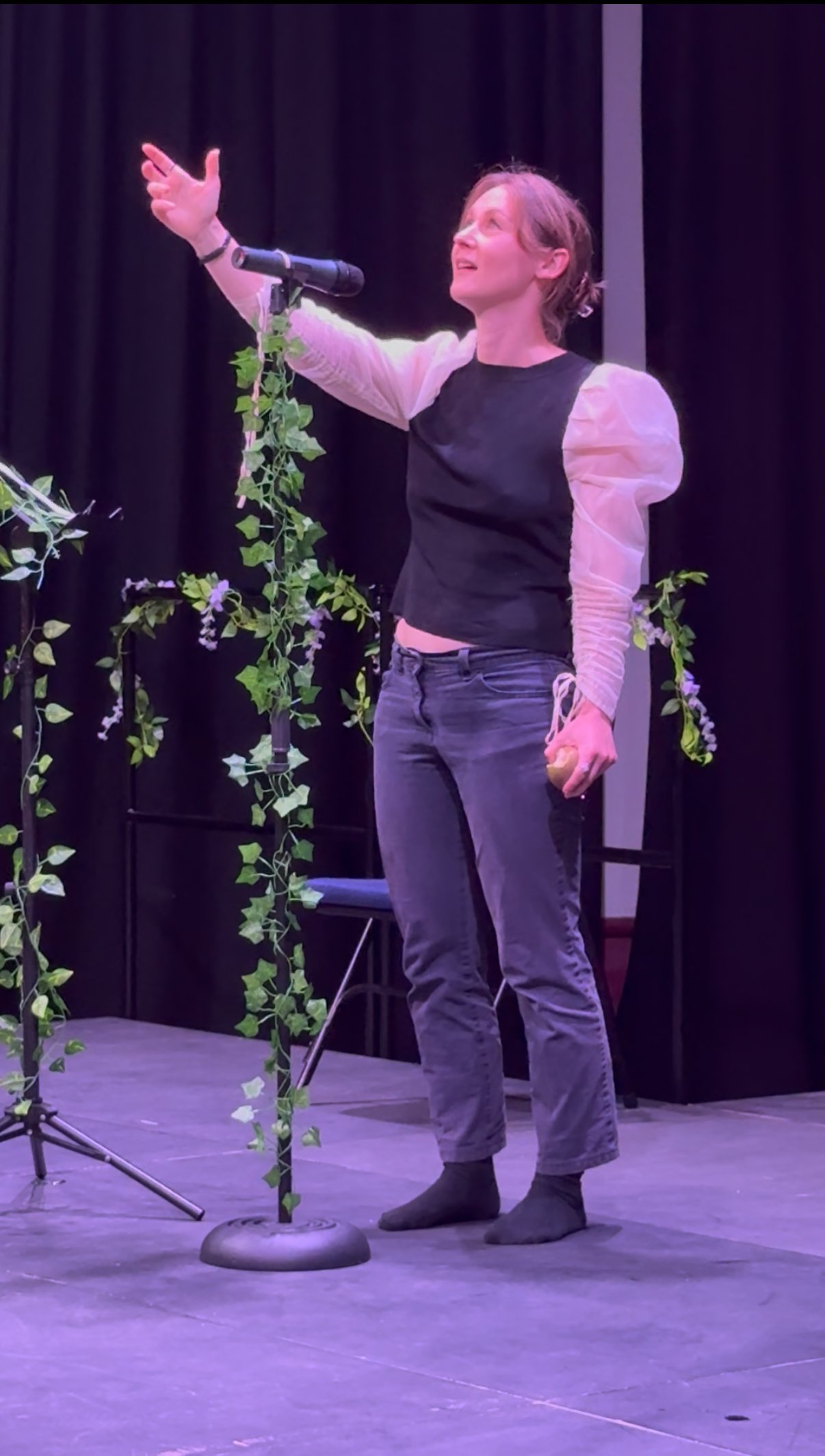 A woman standing on a stage, dressed in a black shirt and gray pants, reaching out with her right arm, with a microphone on a stand decorated with green ivy nearby, in front of a black curtain.
