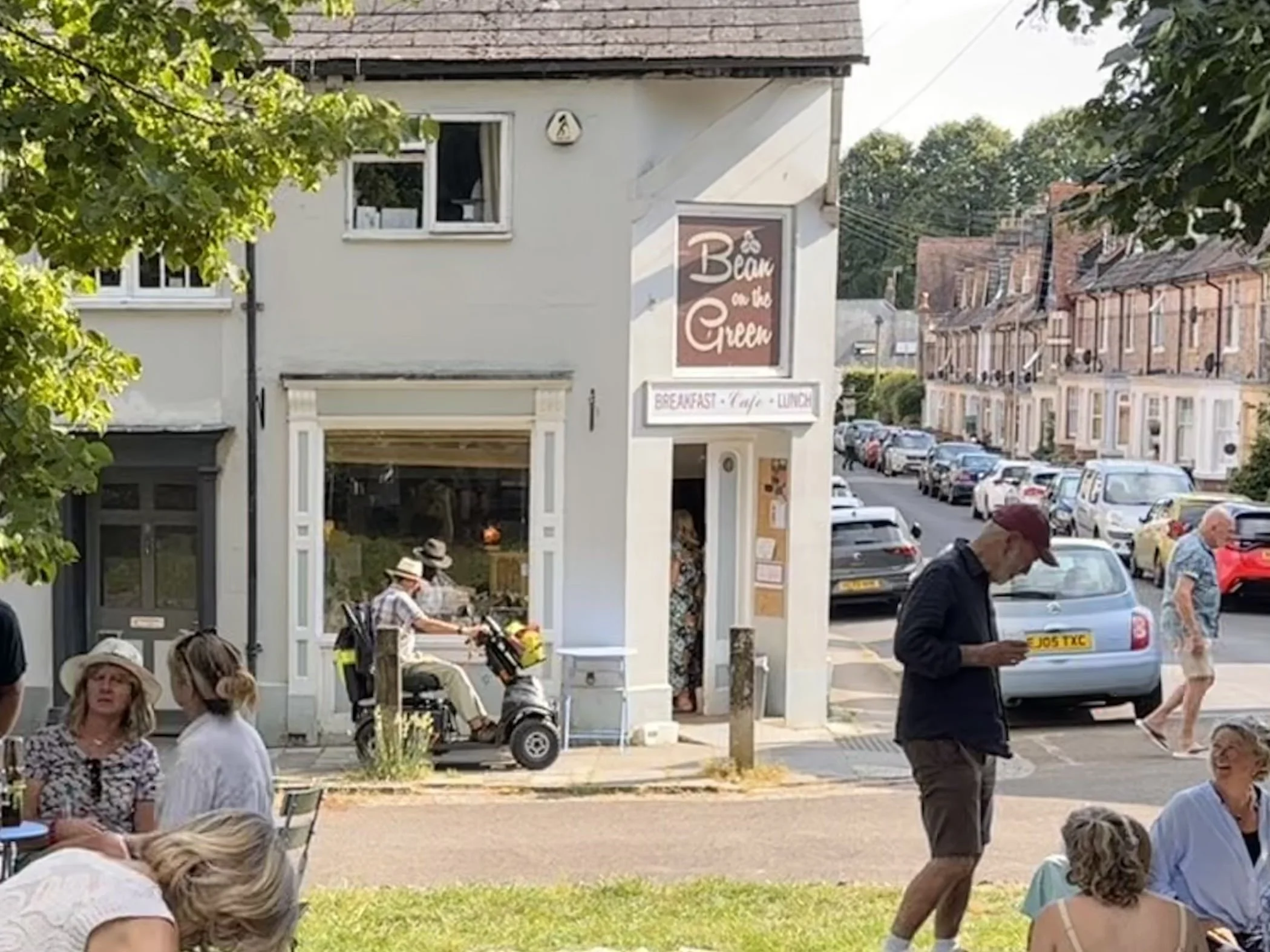 People sitting and standing on a sidewalk in front of a row of houses, with some parked cars in the street. A building with a sign reading 'Bean on the Green' is visible.