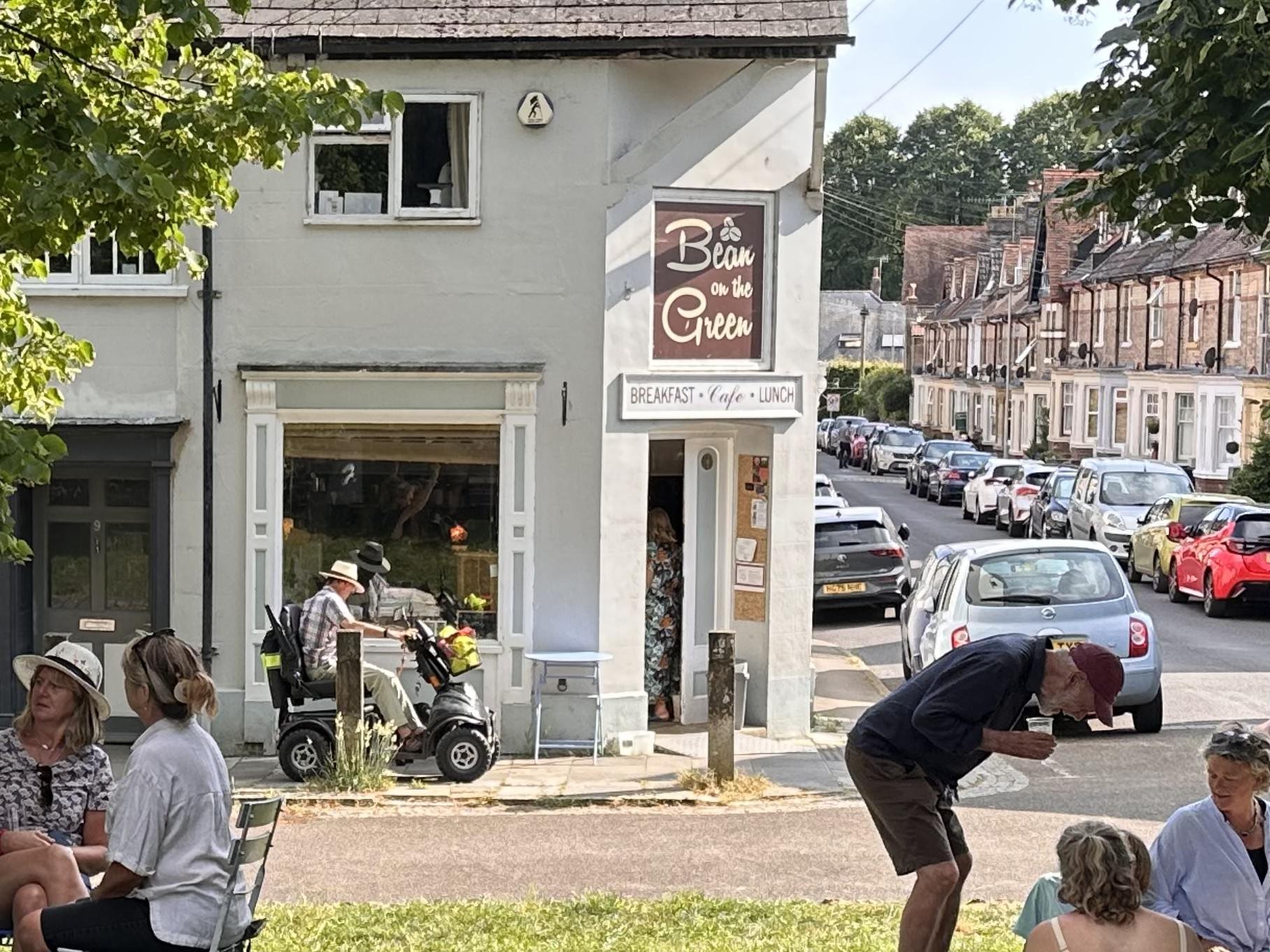 People sitting and talking outside a cafe on a sunny day, with a white building that has a sign reading 'Bean on the Green' and a street with parked cars in the background.