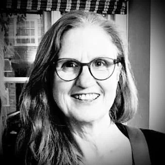 Black and white photo of a woman with long hair, wearing glasses, smiling, indoors with a window in the background.