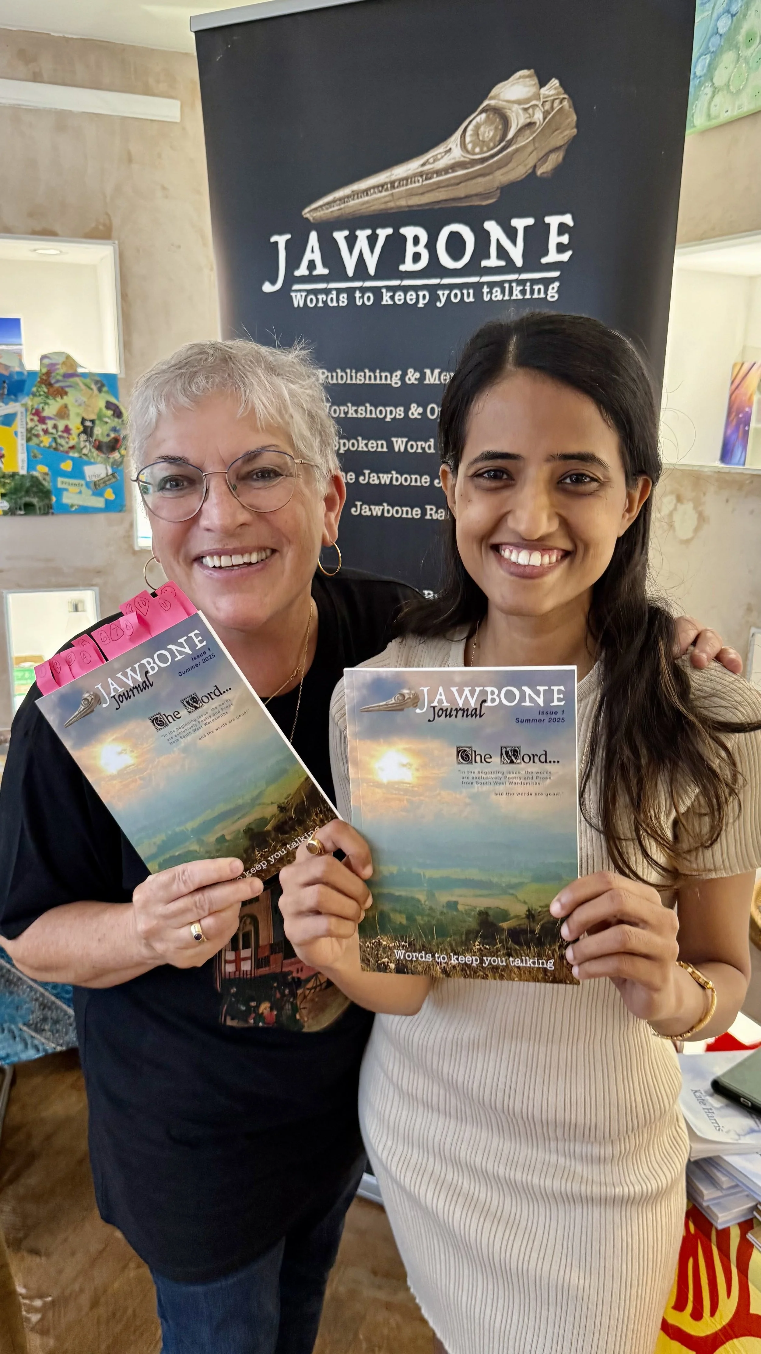 Two women smiling and holding magazines titled 'JAWBONE Journal', standing in front of a black banner with the same title and a dinosaur skull illustration. The banner includes text about publishing, workshops, and spoken words.