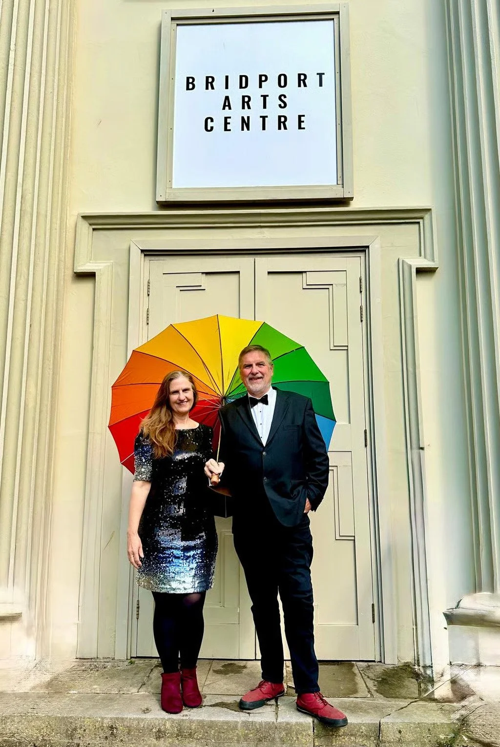 Two people dressed in formal attire standing in front of a door outside the Bridport Arts Centre, holding a colorful rainbow umbrella.