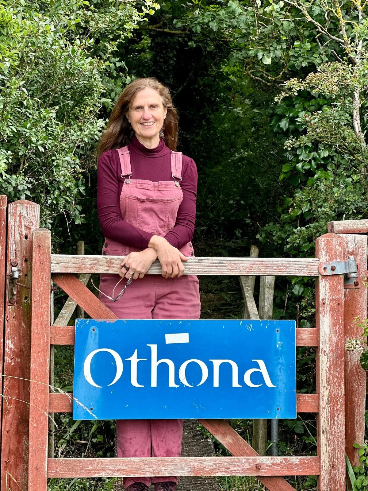 A woman standing behind a wooden gate with a blue sign that says 'Othona'. She is smiling and wearing a maroon turtleneck and maroon overalls. There is green foliage around her.