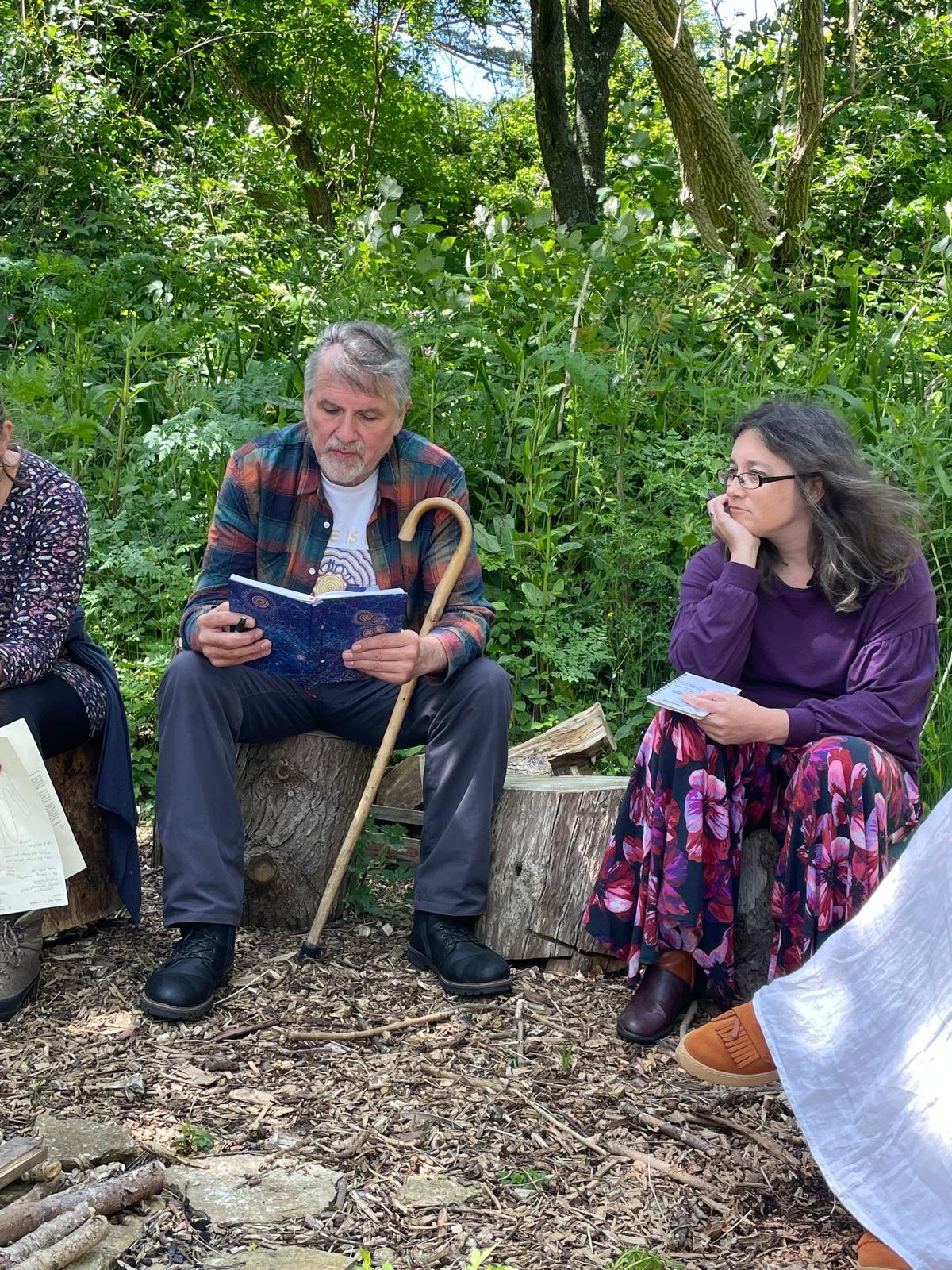 Group of people sitting outdoors on tree stumps in a green forest, engaged in reading or writing, with one man holding a book and a walking stick, and a woman looking on attentively.