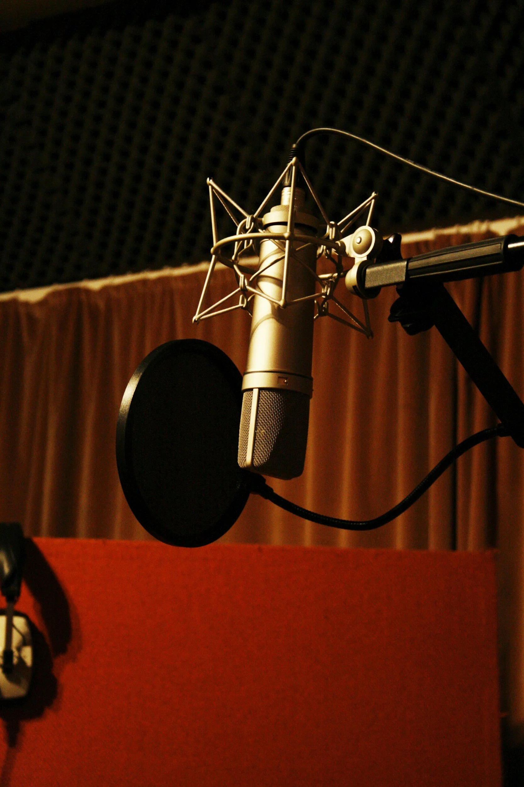 A professional studio microphone with a pop filter in front of a black soundproofing wall and wooden panels with a red surface in the foreground.