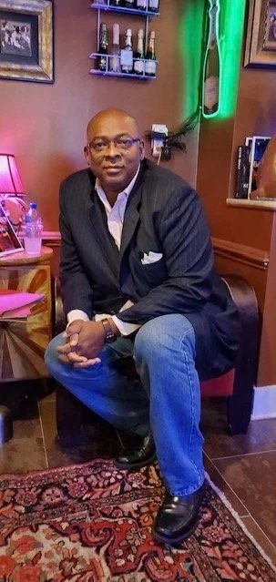 A man sitting on a chair in a restaurant or bar, wearing a dark blazer, light shirt, and blue jeans, with bottles of beverages on a shelf behind him and framed pictures on the wall.