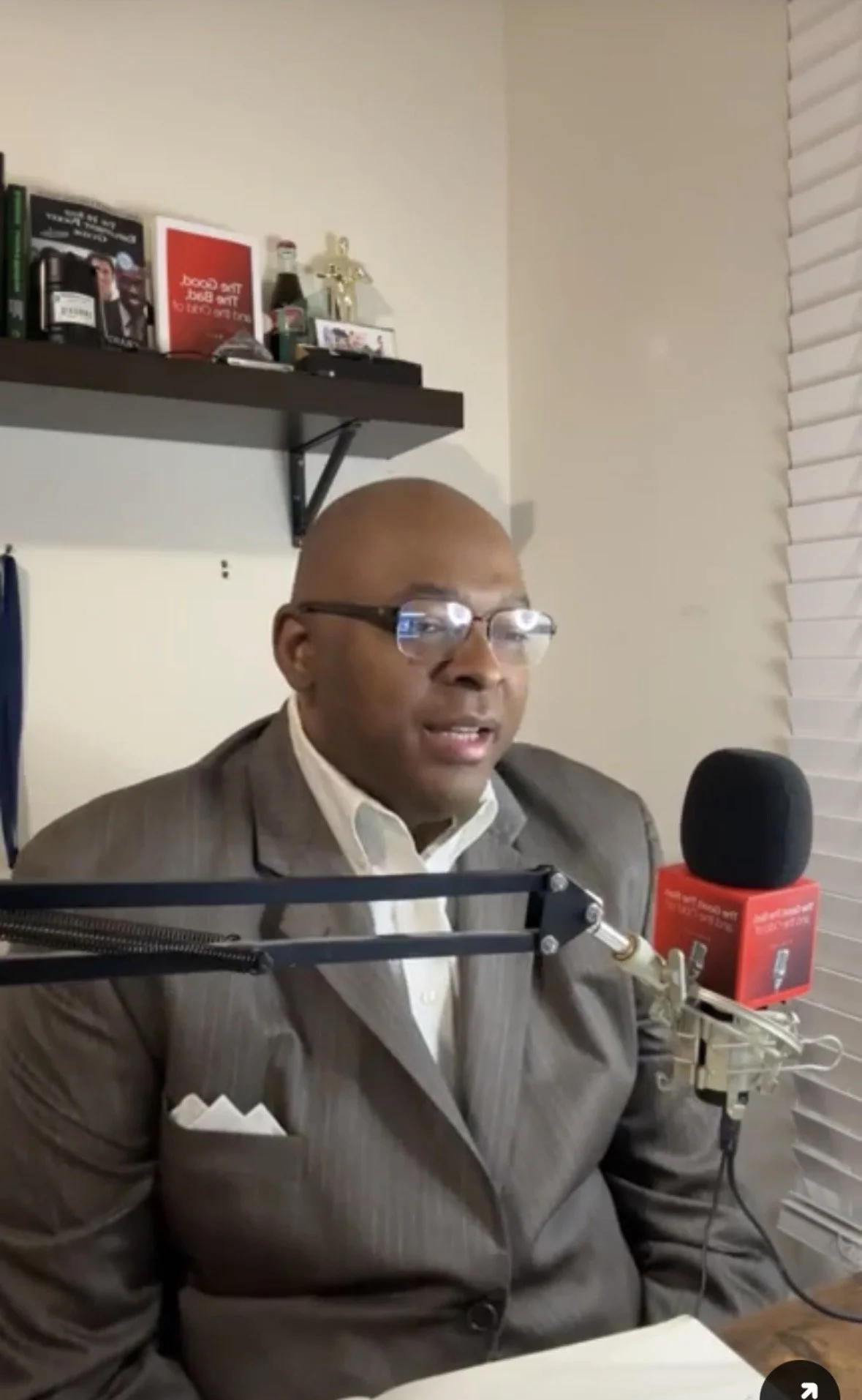A man wearing glasses and a suit speaking into a microphone in a studio setting.