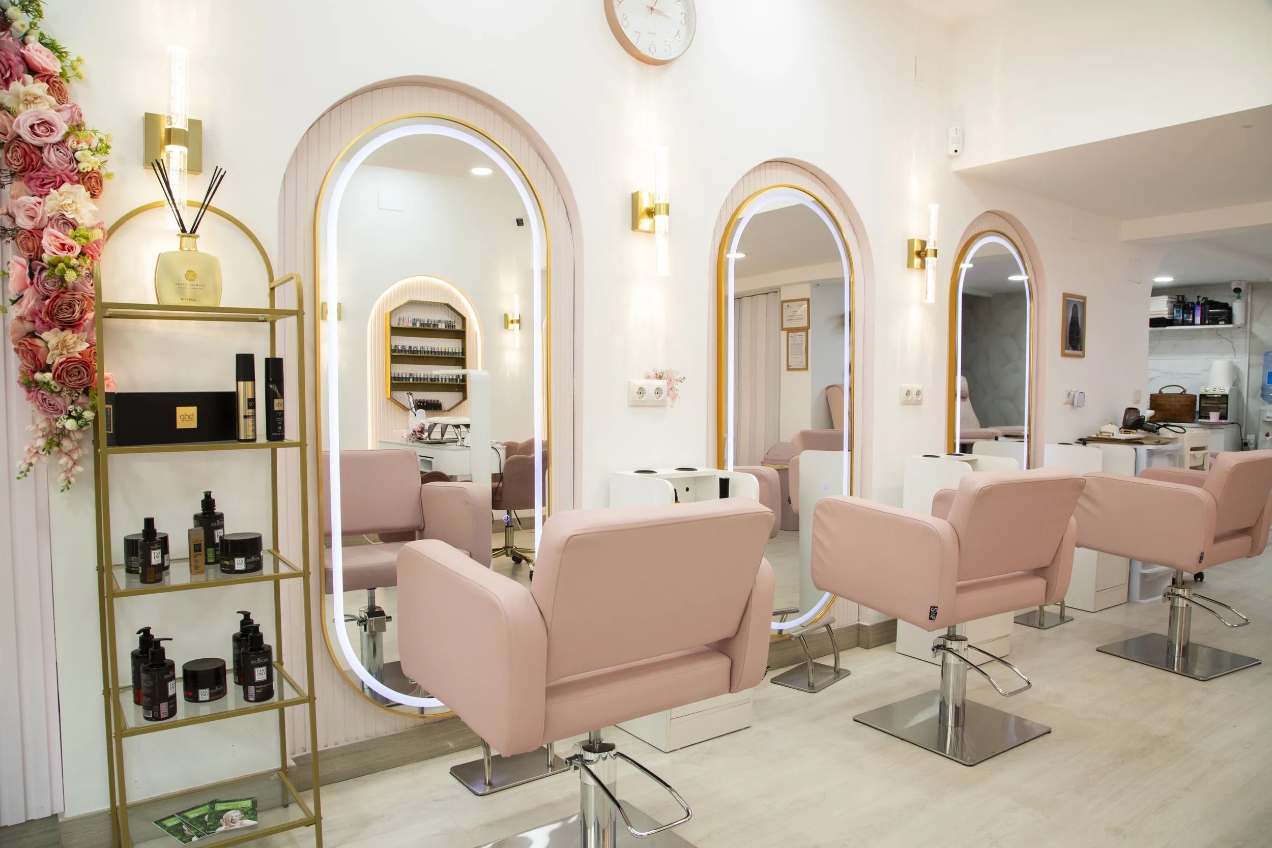 Interior de un salón de belleza con sillas rosadas, espejos con marcos dorados, estantes con productos y decoración floral en una esquina.