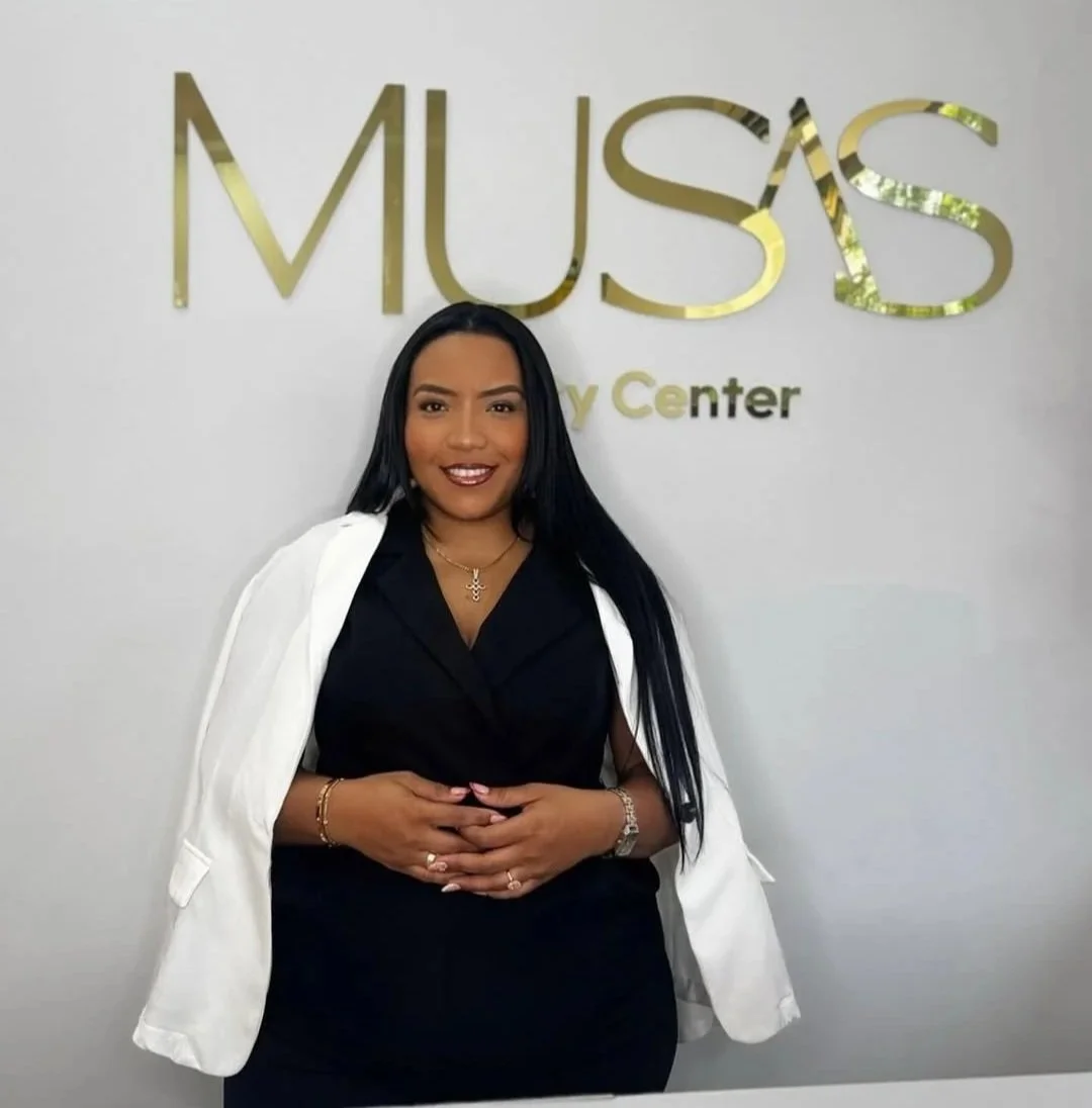 Mujer sonriendo frente a una pared con el cartel de 'Musas Beauty Center', vistiendo ropa profesional y accesorios dorados.