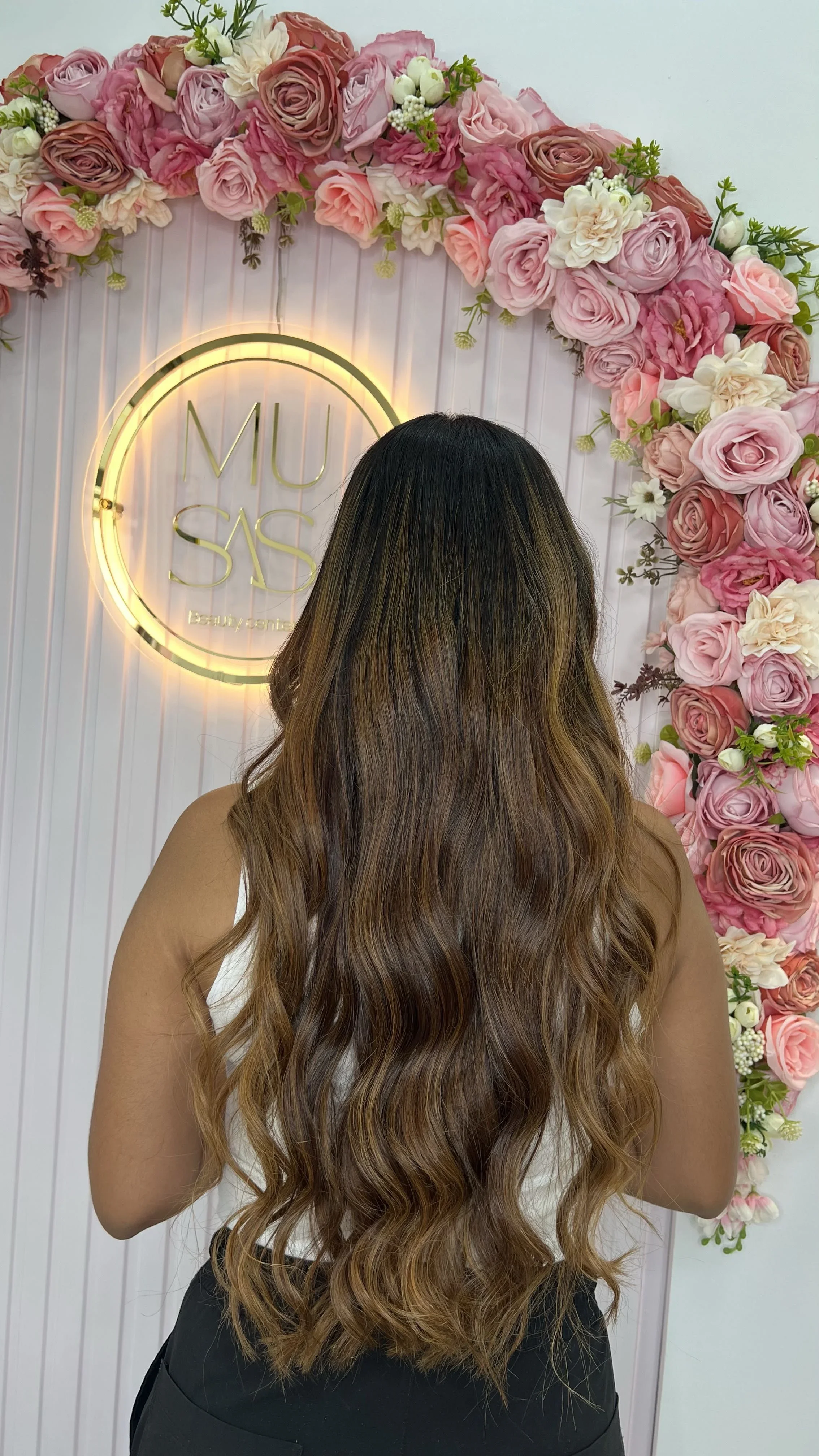 Persona con cabello largo, castaño y con ondas, de espaldas, frente a fondo decorado con flores y un cartel que dice 'MU SAS'.