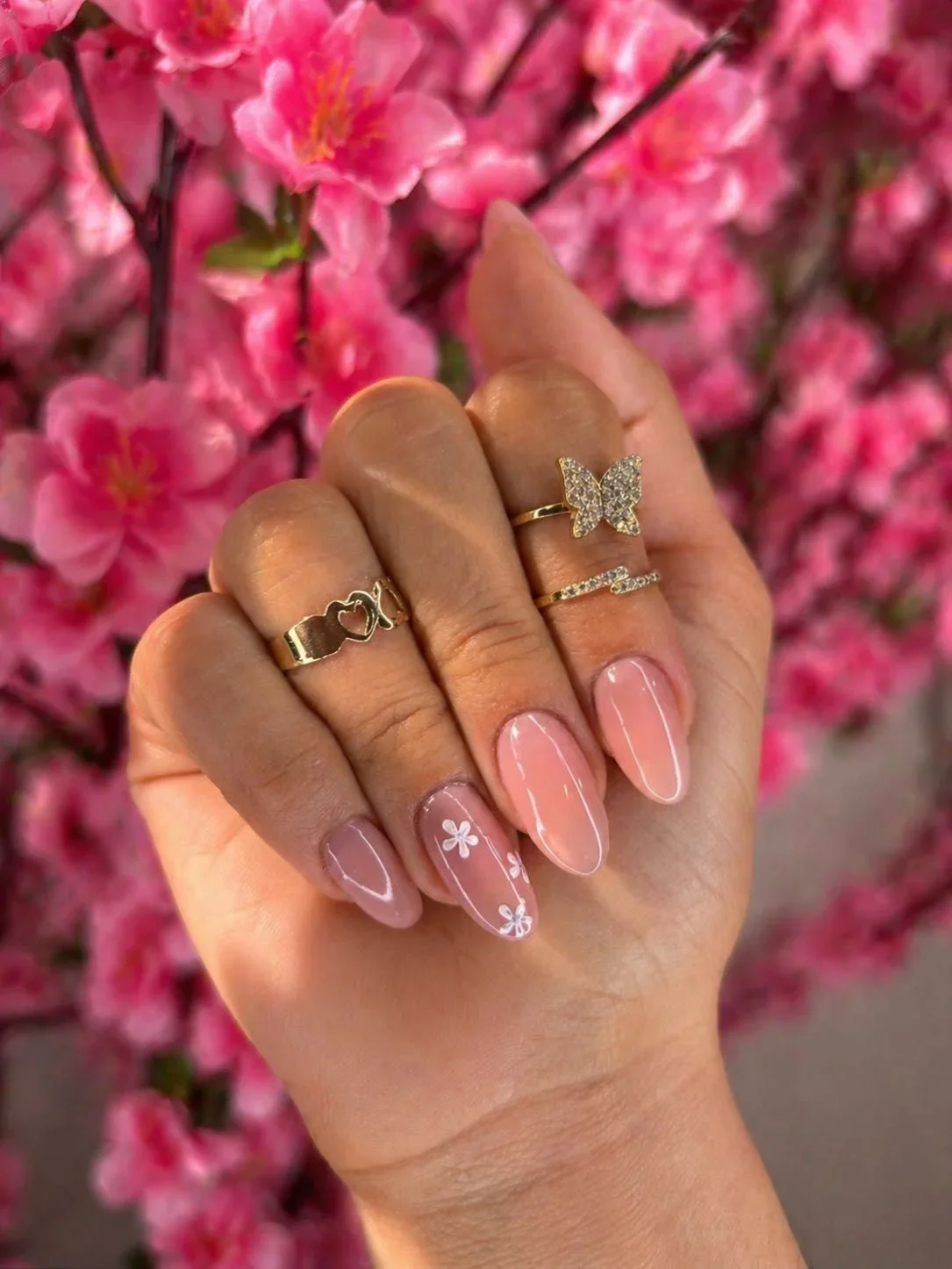 Mano con uñas con diseño de flores, usando varios anillos, uno con mariposa y otros con corazones, en un fondo de flores rosadas.