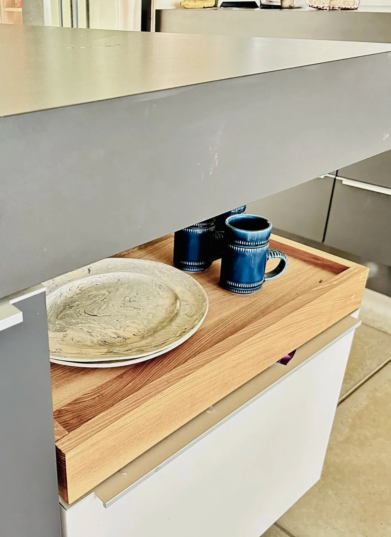 A beige and gray countertop with a wooden tray holding two dark blue cups, a beige decorative plate with a swirling pattern, and some pieces of cake in the background.