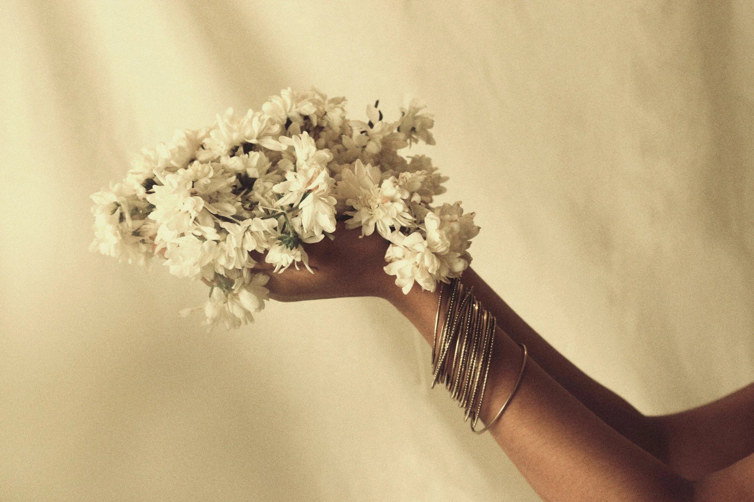 A person holding a bouquet of white flowers with a neutral background, wearing multiple silver and gold bracelets on their wrist.