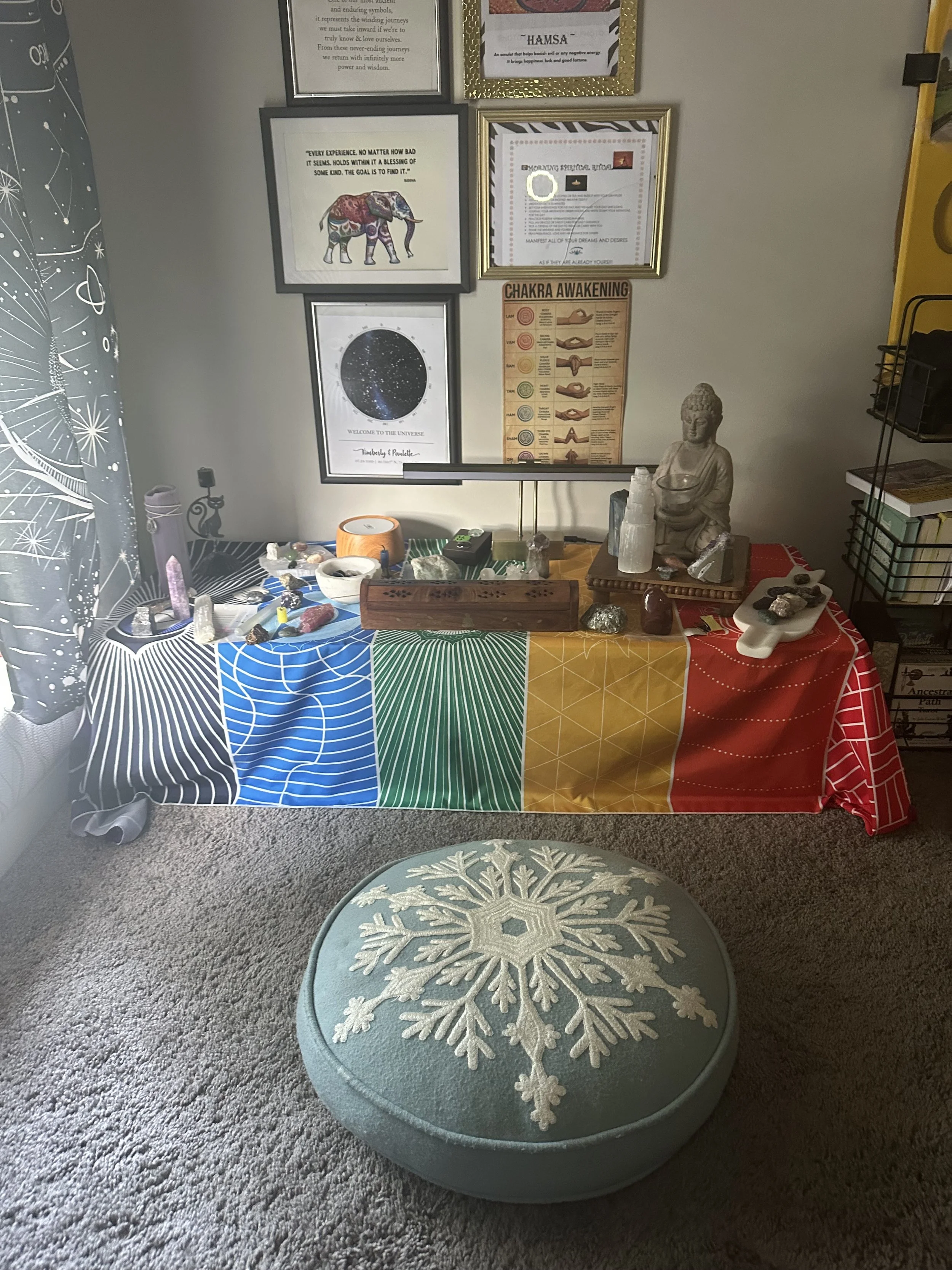 A table covered with a colorful cloth with star, planetary, and rainbow patterns, holding various stones, crystals, and objects. Behind the table, a collection of framed quotes and artistic posters on a wall. To the right, a small statue of Buddha, and a small tray with stones. A round ottoman with a large snowflake design on the top is in the foreground, and part of a curtain with star constellations on the left.