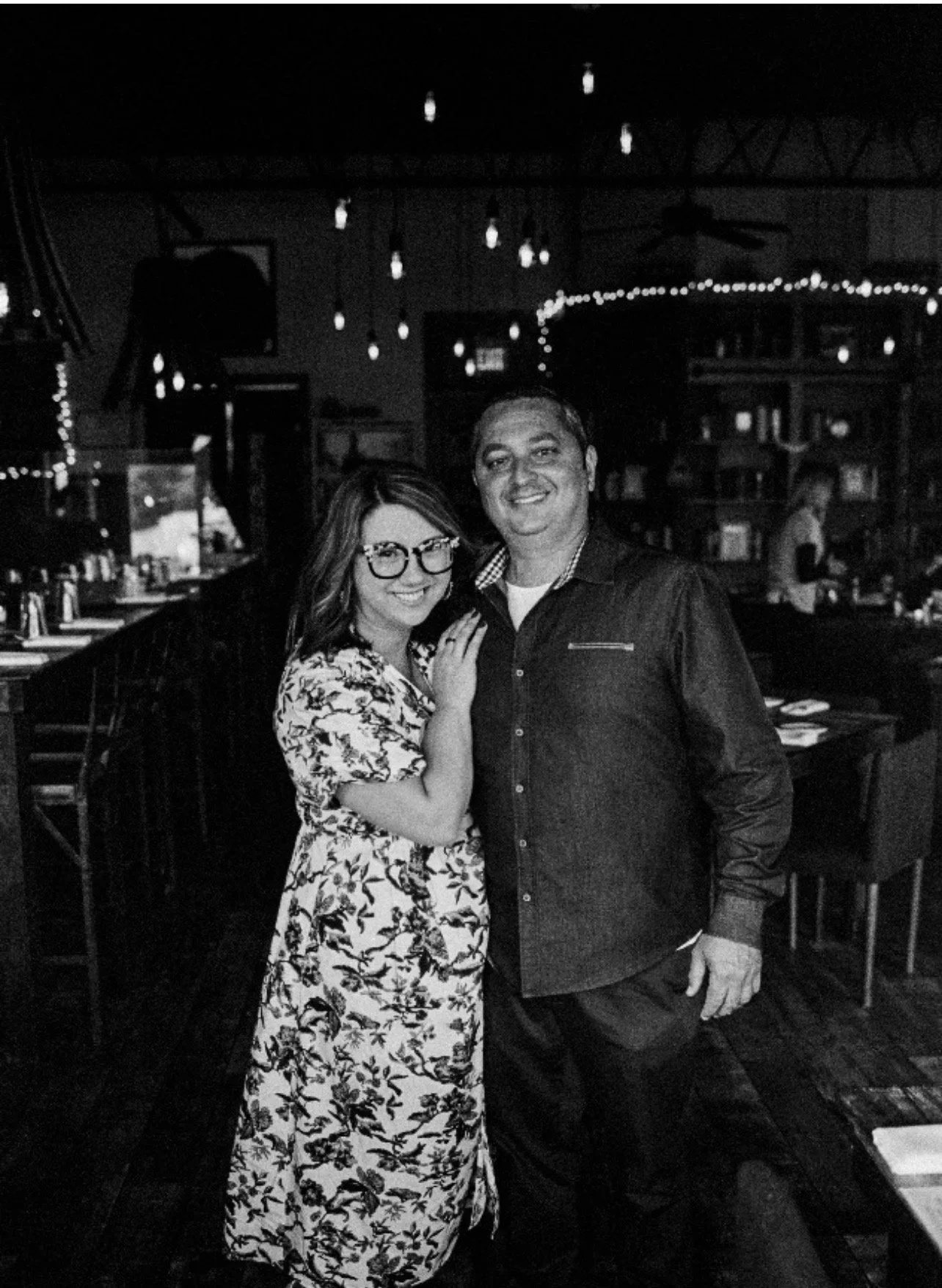 A black-and-white photo of a smiling woman wearing glasses and a floral dress, standing next to a man in a dark shirt, inside a dimly lit restaurant or bar with hanging string lights and shelves behind them.