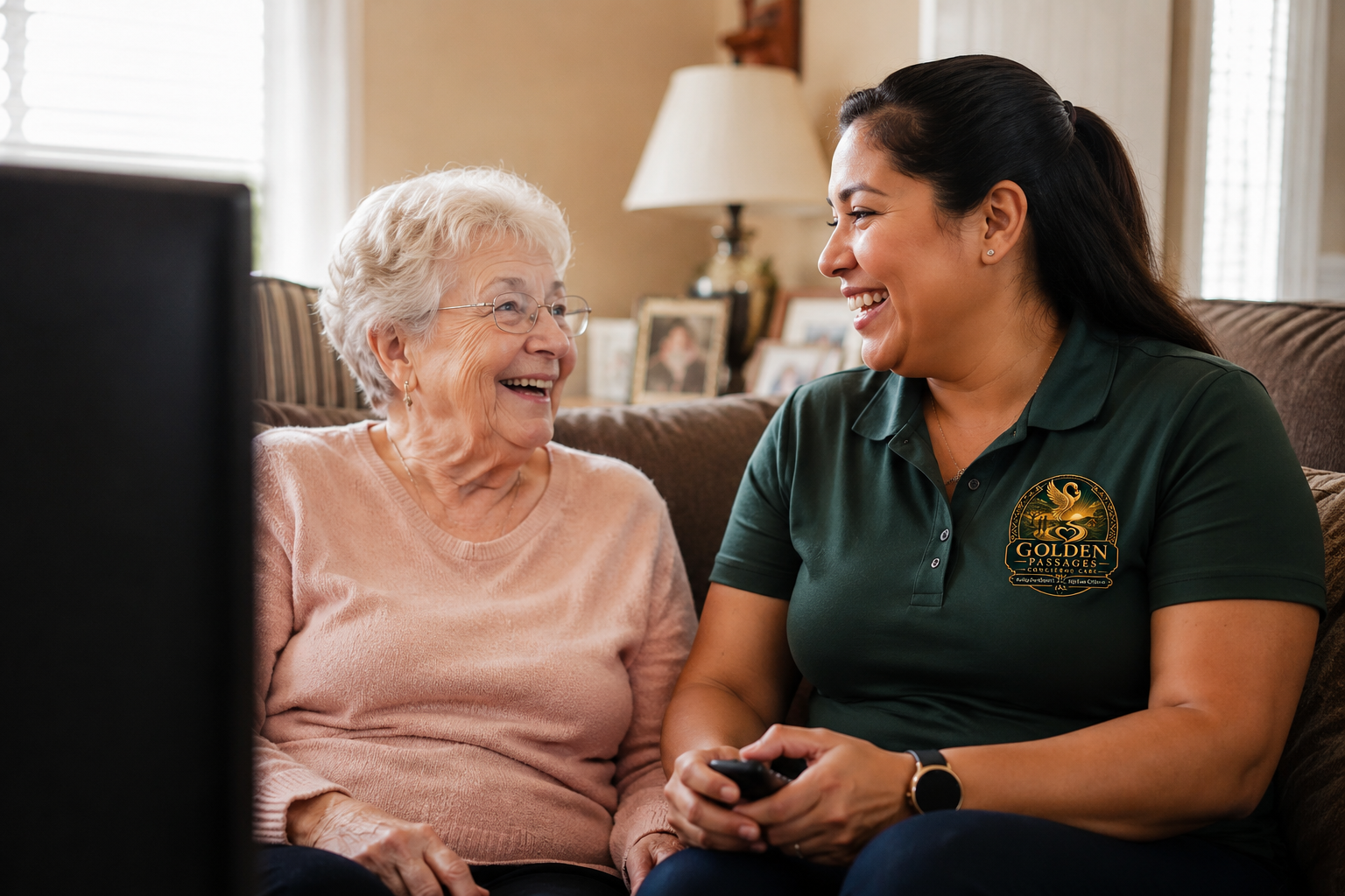 An elderly woman with white hair and glasses sitting on a couch, smiling at a young woman with dark hair in a ponytail, wearing a green shirt with a logo, holding a remote control, inside a cozy living room.