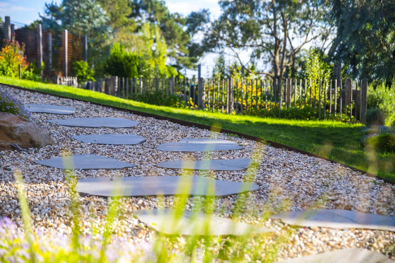 A garden pathway with round stepping stones on gravel, grass, trees, and a wooden fence in the background, under sunny weather.