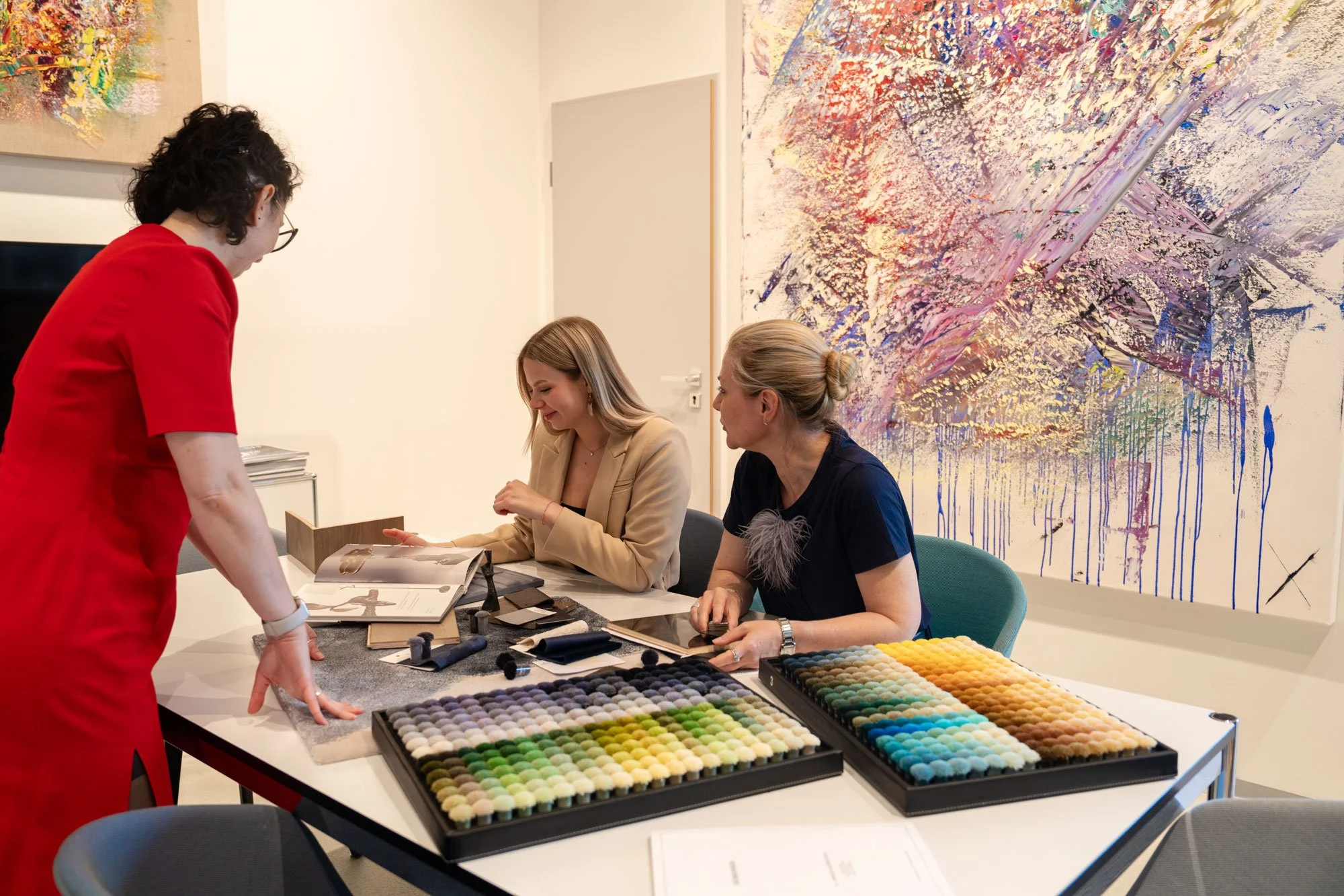 Drei Frauen sitzen an einem Tisch in einem Kunstmuseum oder einer Galerie und betrachten Stoffproben und Farbpaletten. Eine Frau in rotem Mantel steht, während die beiden anderen Frauen die Stoff- und Farbproben anschauen. Im Hintergrund gibt es bunte abstrakte Kunstwerke an der Wand.