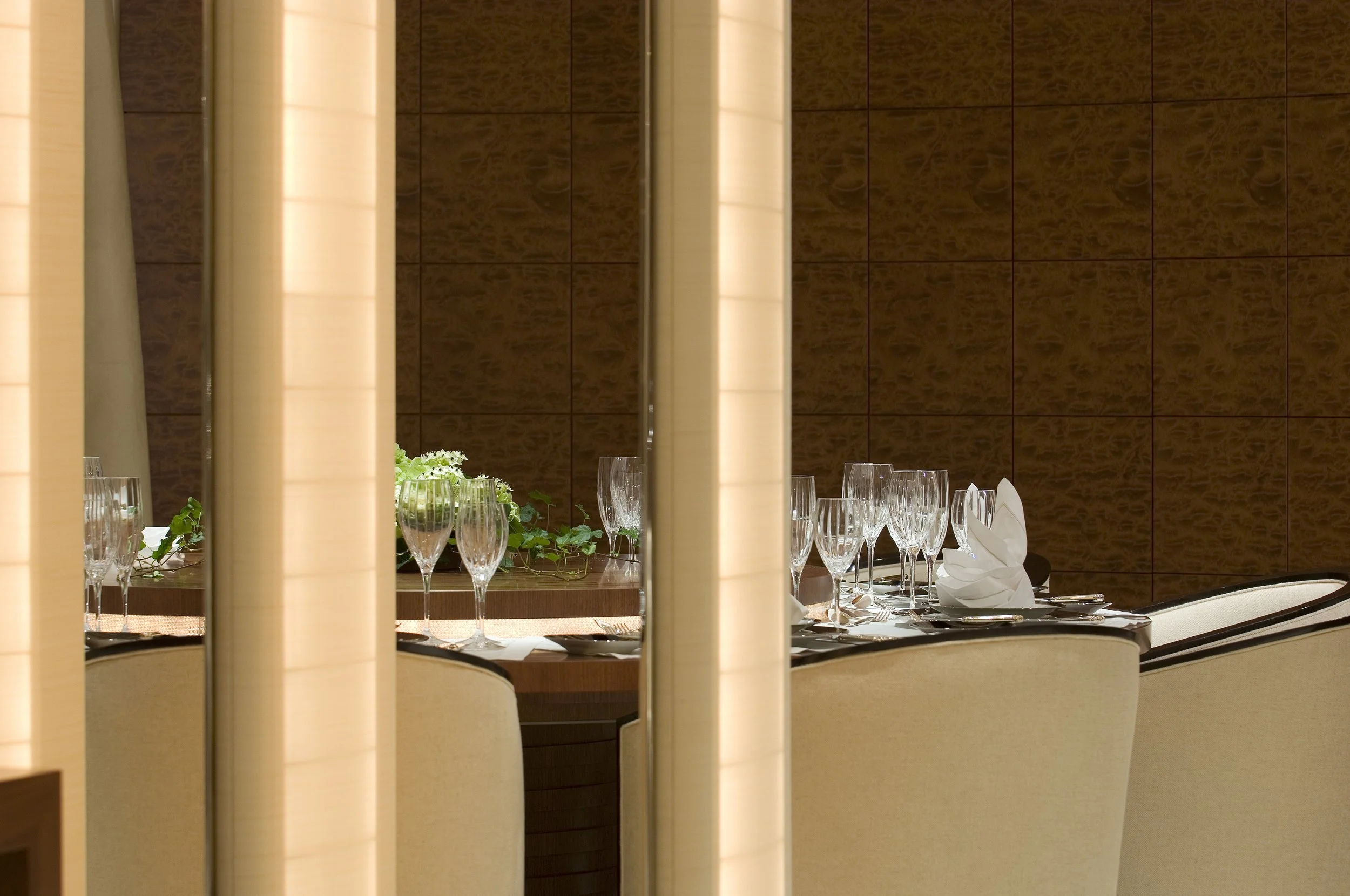 Elegant Empfangstresen mit Wein- und Wassergläser, Kerzenschein, Blumenteller und Besteck, in einem modernen Bereich mit Holz- und dunklen Wandflächen, teilweise durch weiße Säulen getrennt.