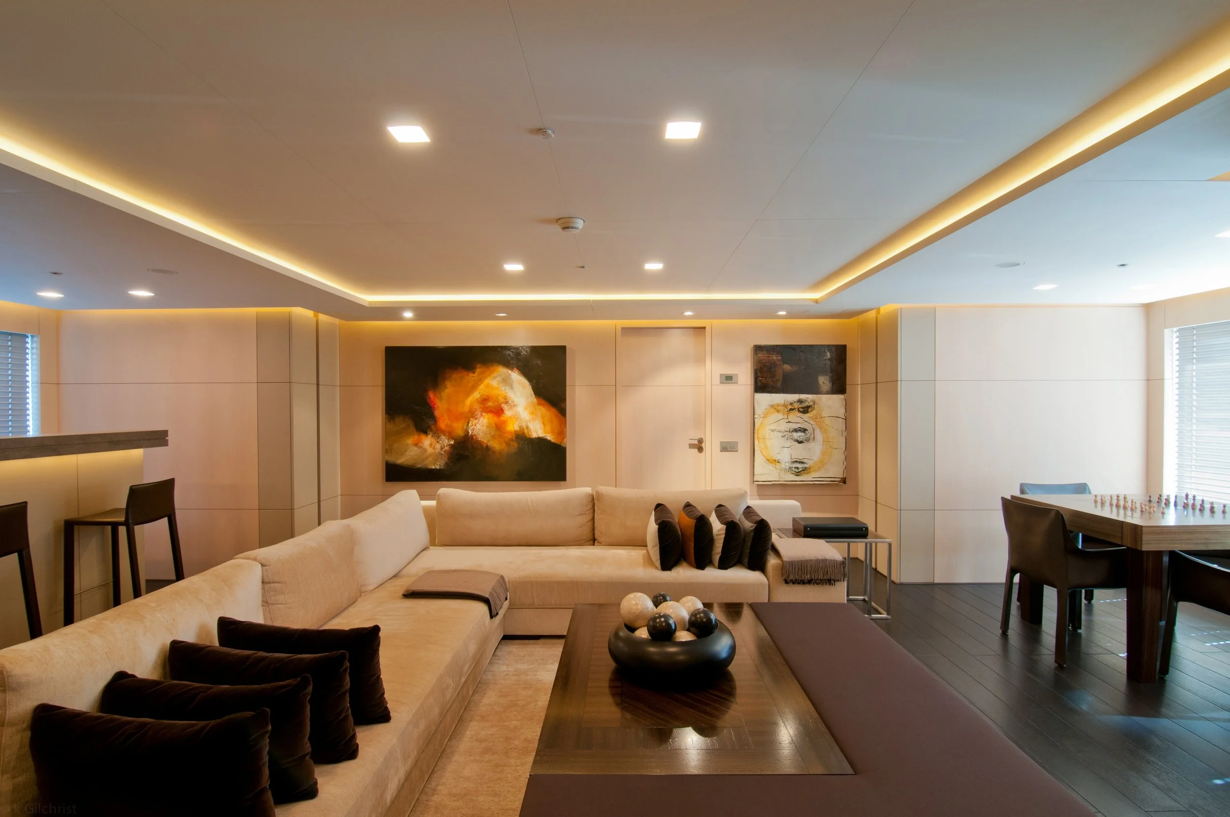 Moderner Wohnraum mit Sofa, Kunstwerken an der Wand, Esstisch und Fenstern mit Jalousien, helle Decke mit eingebauten Lichtleisten.
