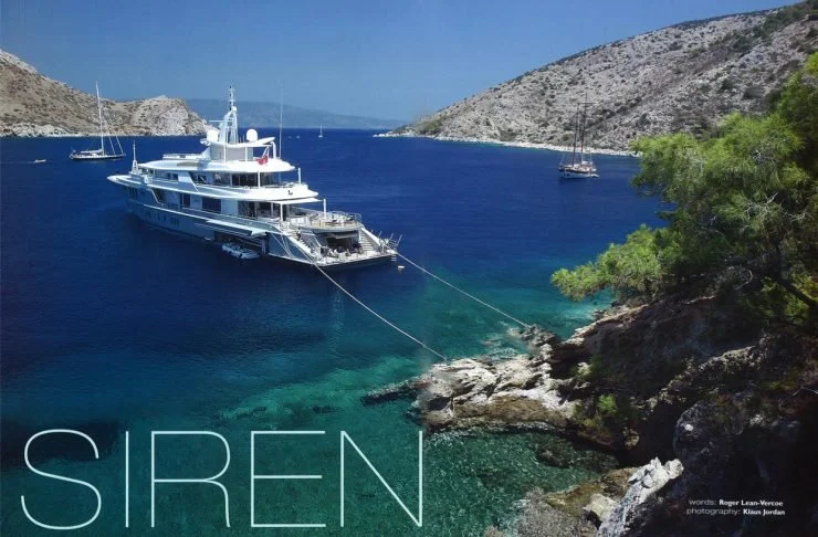 Ein großes weißes Yacht liegt in einer Bucht mit klarem blauen Wasser, umgeben von felsigen Hügeln und Bäumen, mit kleineren Segelbooten im Hintergrund.