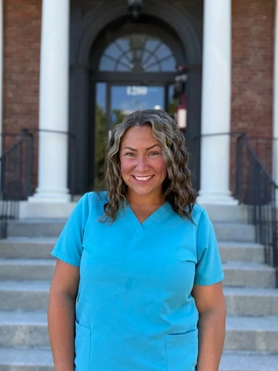 A woman smiling and wearing a blue medical scrub top standing outside a brick building with a large arched window and stairs.