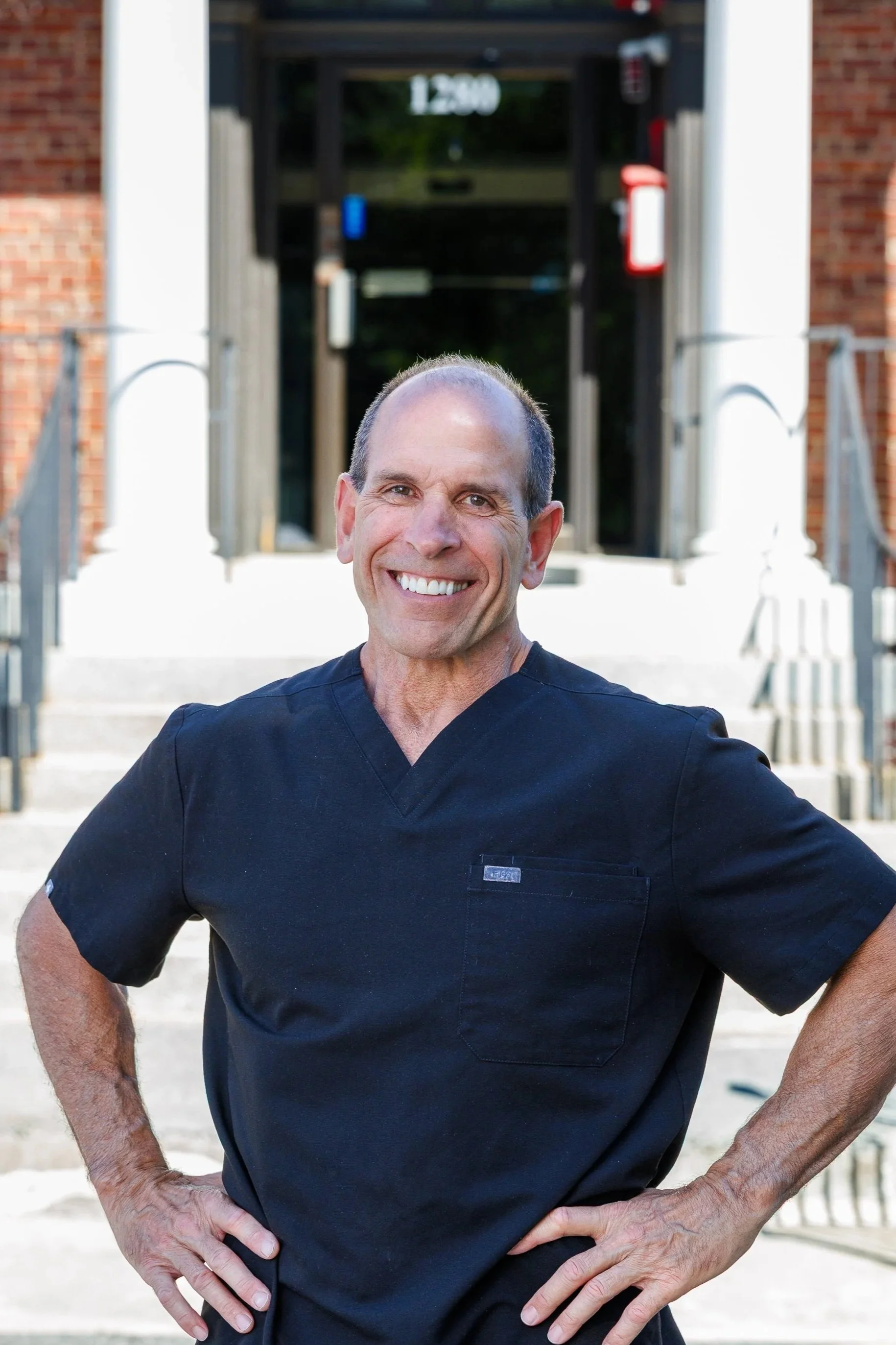 A smiling man with short hair and a black medical scrub top standing outdoors with arms crossed, blurred brick and metal structure in background.
