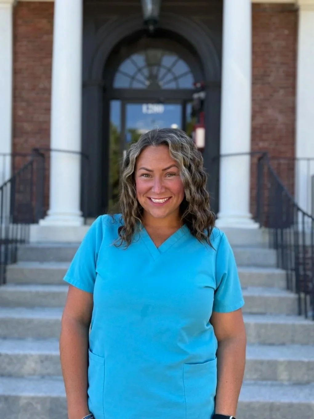A woman in blue scrubs standing outside a building with stairs, brick walls, white columns, black railings, and an arched window.