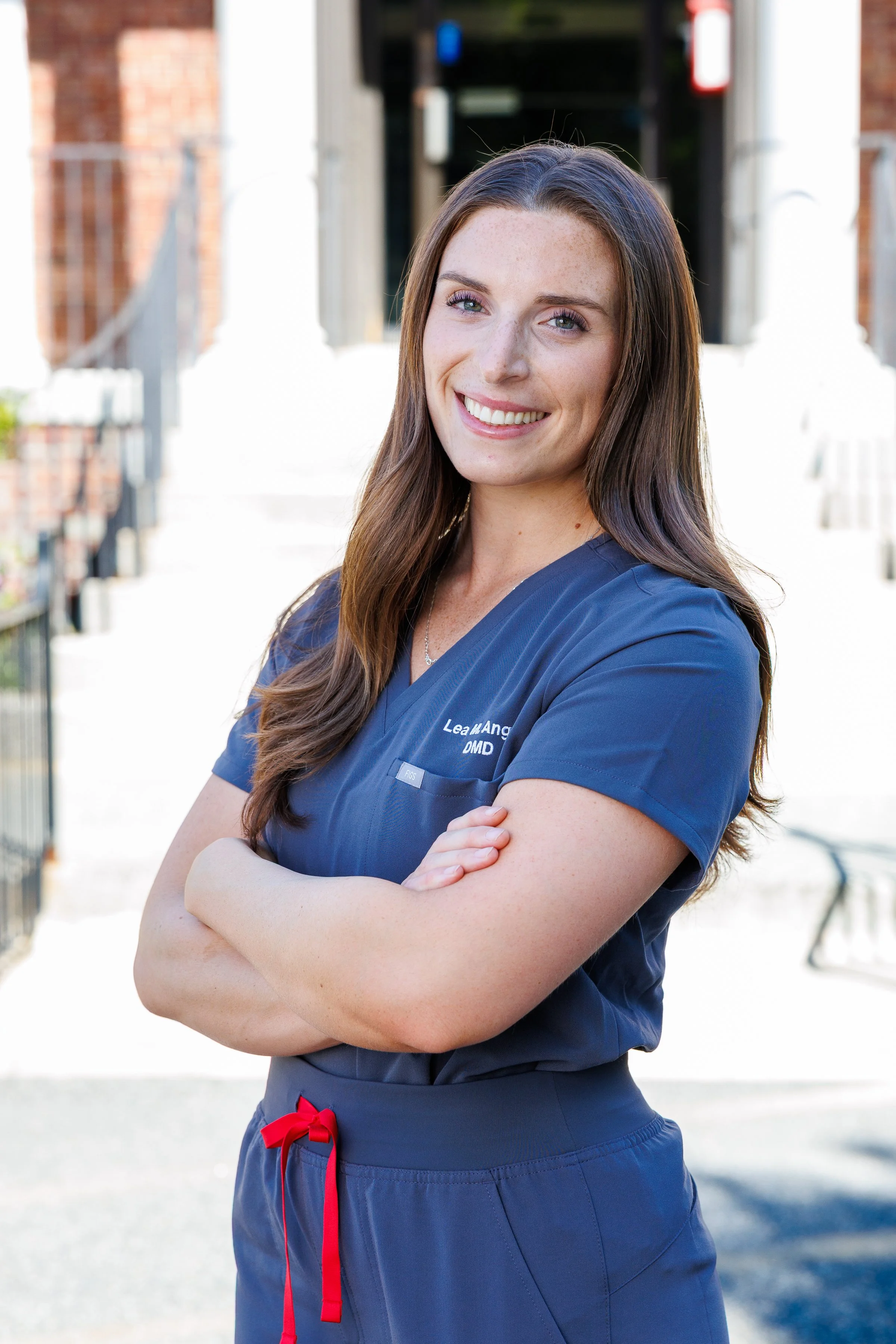 A young female medical professional with long brown hair and blue eyes smiling outdoors, wearing blue scrubs with her arms crossed
