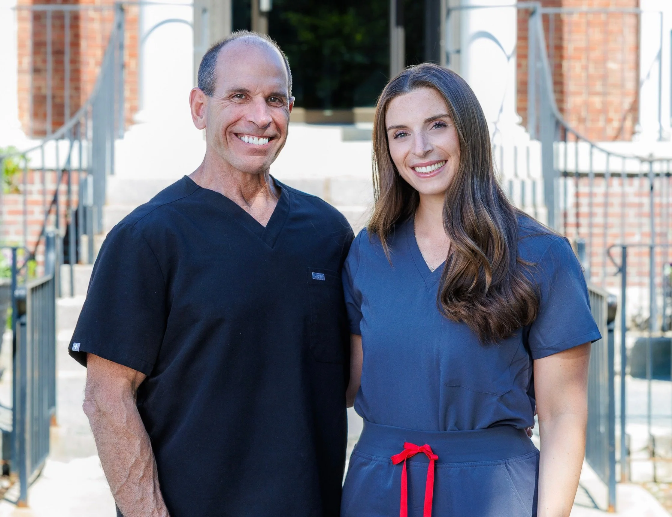 A man and a woman, both wearing navy blue scrubs, standing outside on stairs with brick buildings and metal railings, smiling at the camera.