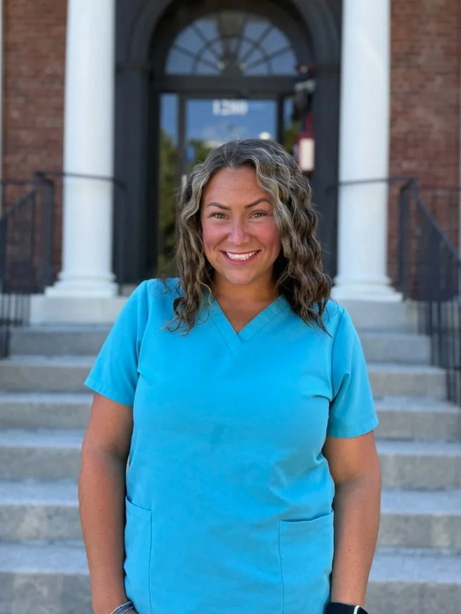 A smiling woman with curly hair wearing blue scrubs standing on front steps of a brick building.