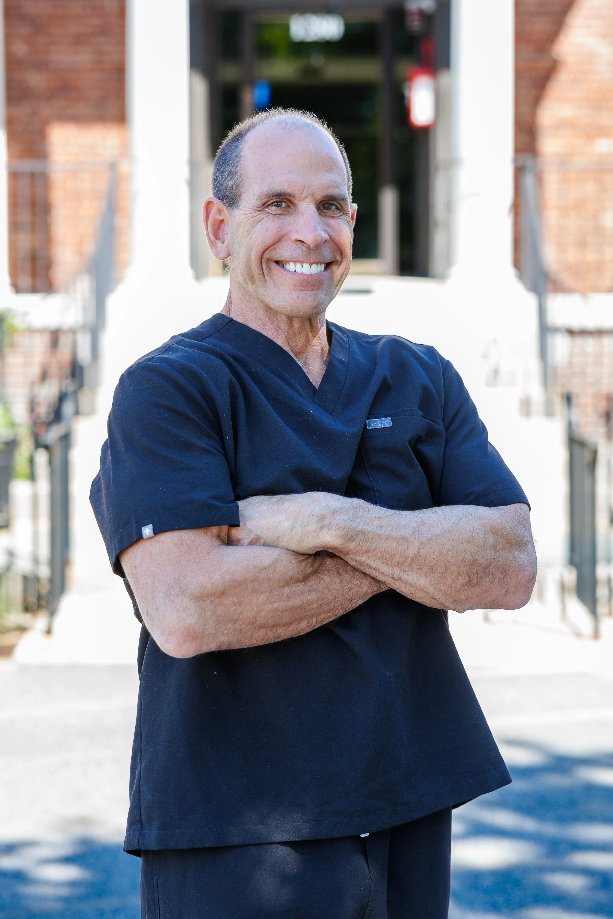 A middle-aged man in a black medical scrub uniform, arms crossed, smiling confidently, standing outdoors in front of a building with brick walls and steps.