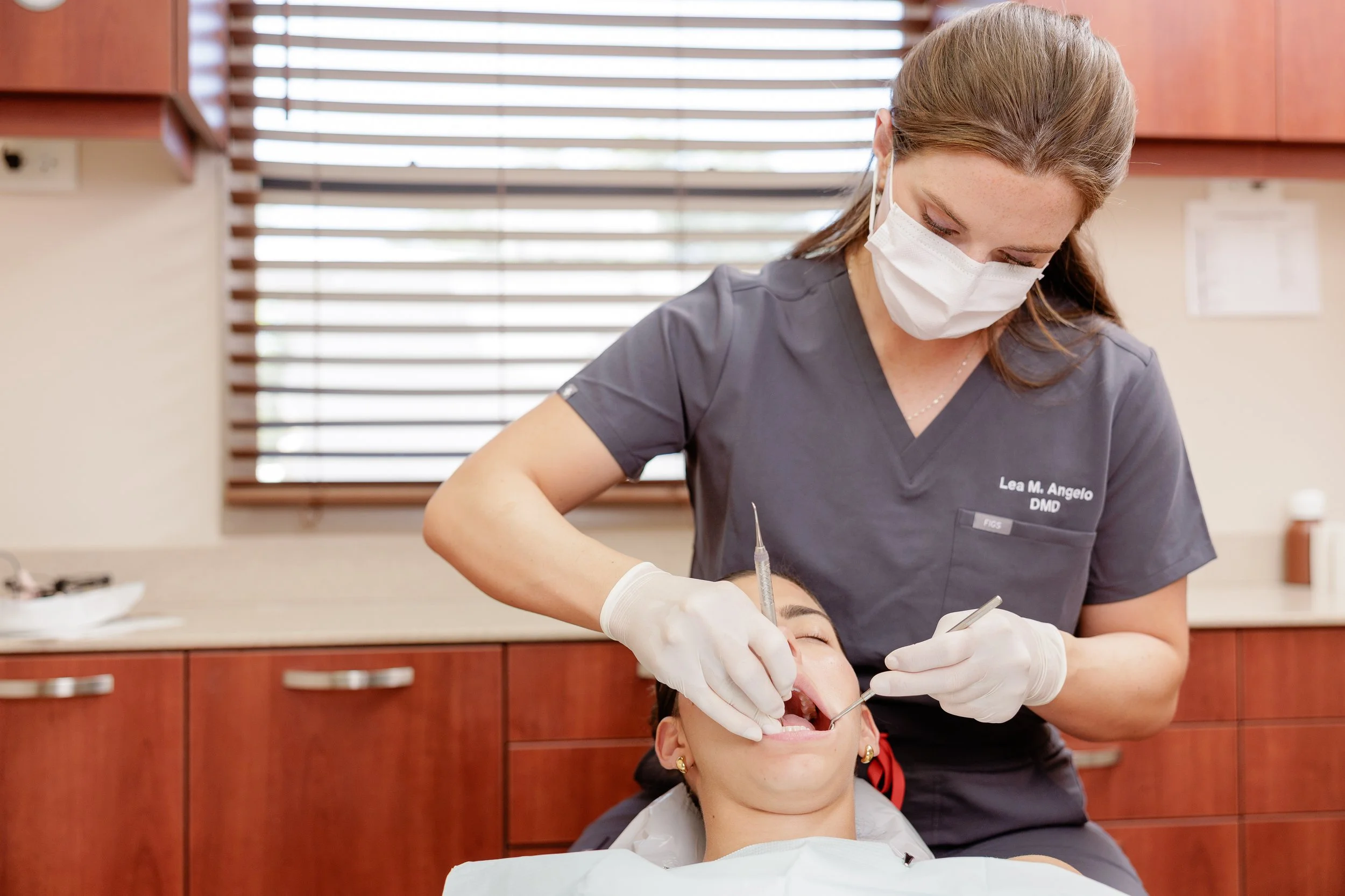 Dentist in gray scrubs and face mask performing dental work on a patient in a dental office.