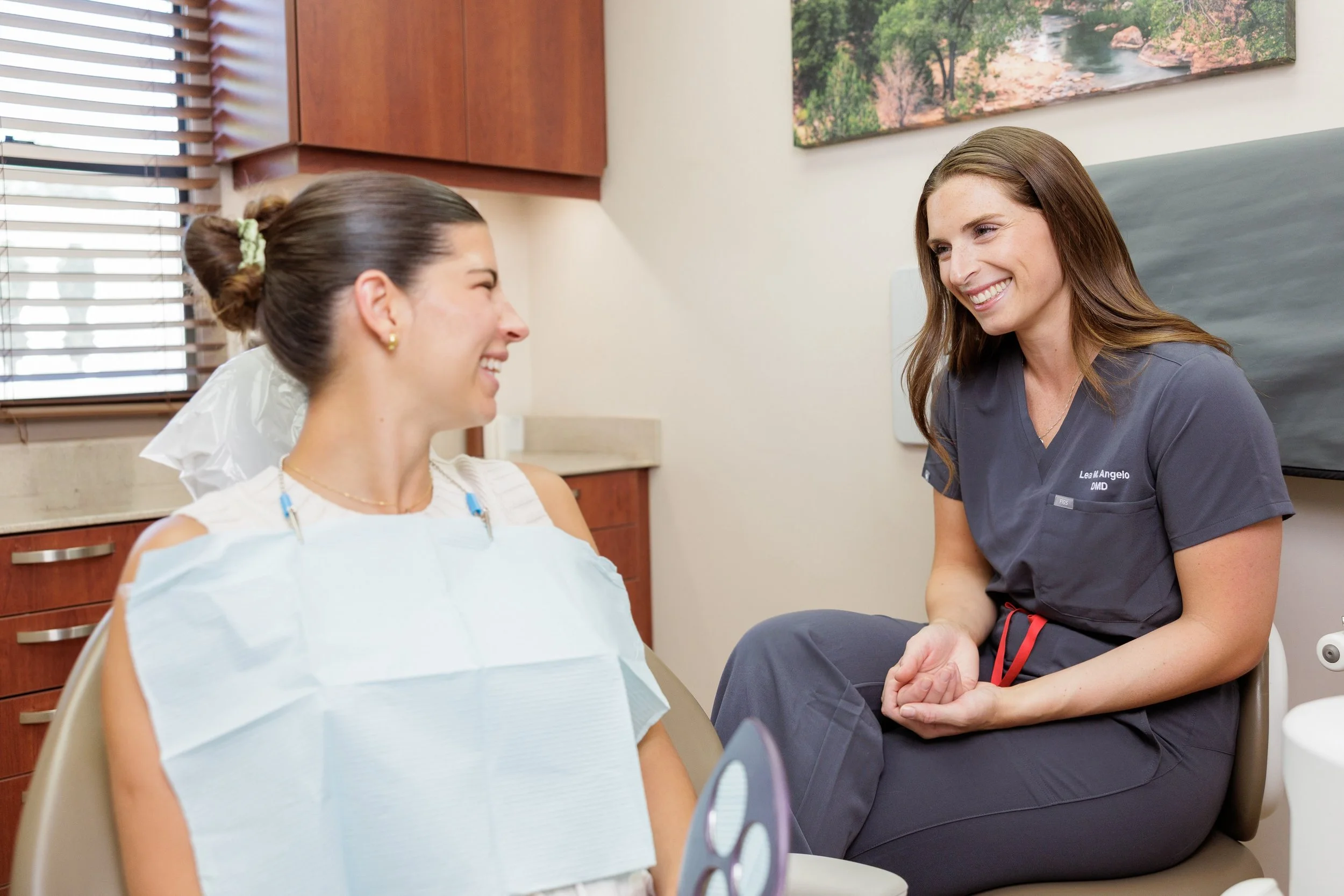 A woman in a hospital bed smiling at a female healthcare professional, who is sitting beside her and smiling back. The healthcare provider is wearing scrubs with a name tag and has long brown hair. The woman in the bed has dark hair tied in a bun, wears earrings, and has a medical gown and a paper bib. The room has wooden cabinets, a window with blinds, and a nature-themed picture on the wall.
