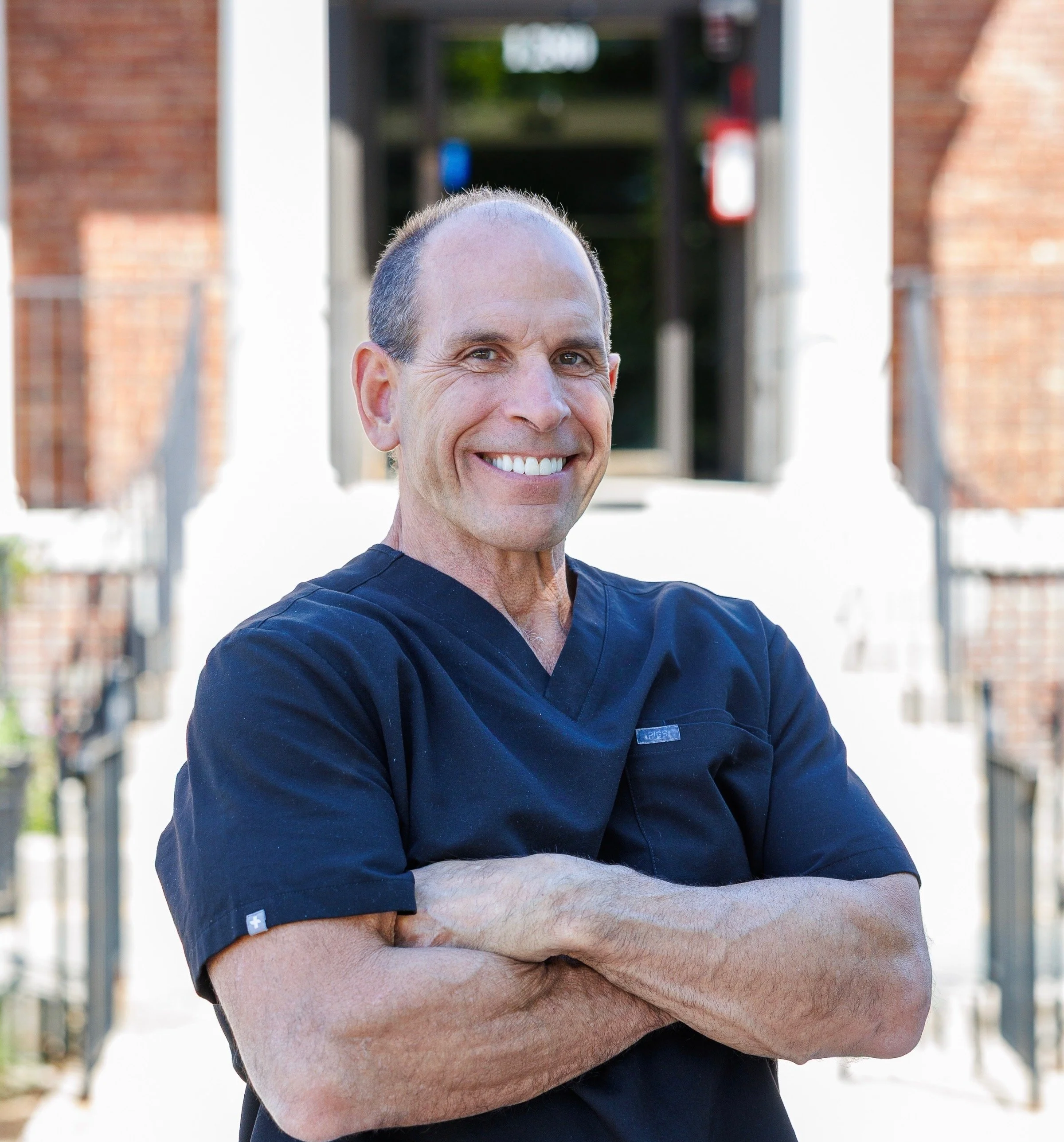 A smiling man with short hair and a black medical scrub top standing outdoors with arms crossed, blurred brick and metal structure in background.