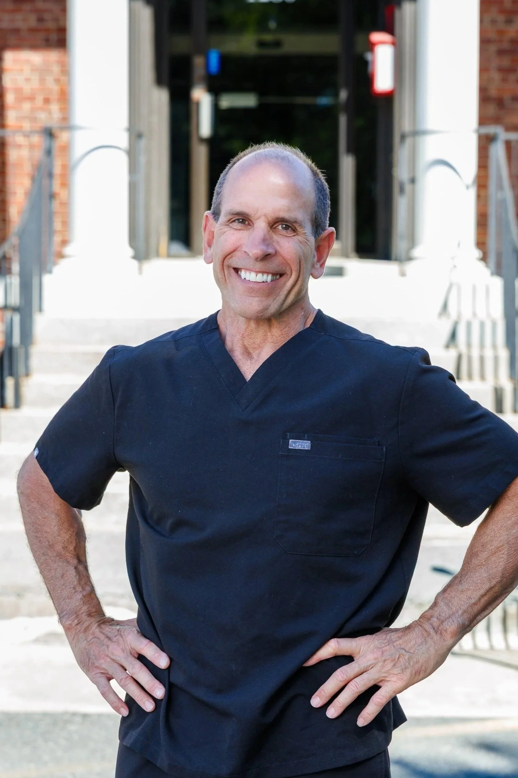 A smiling man in a navy blue medical uniform standing outdoors with hands on hips, in front of the entrance to a building with stairs and brick walls.