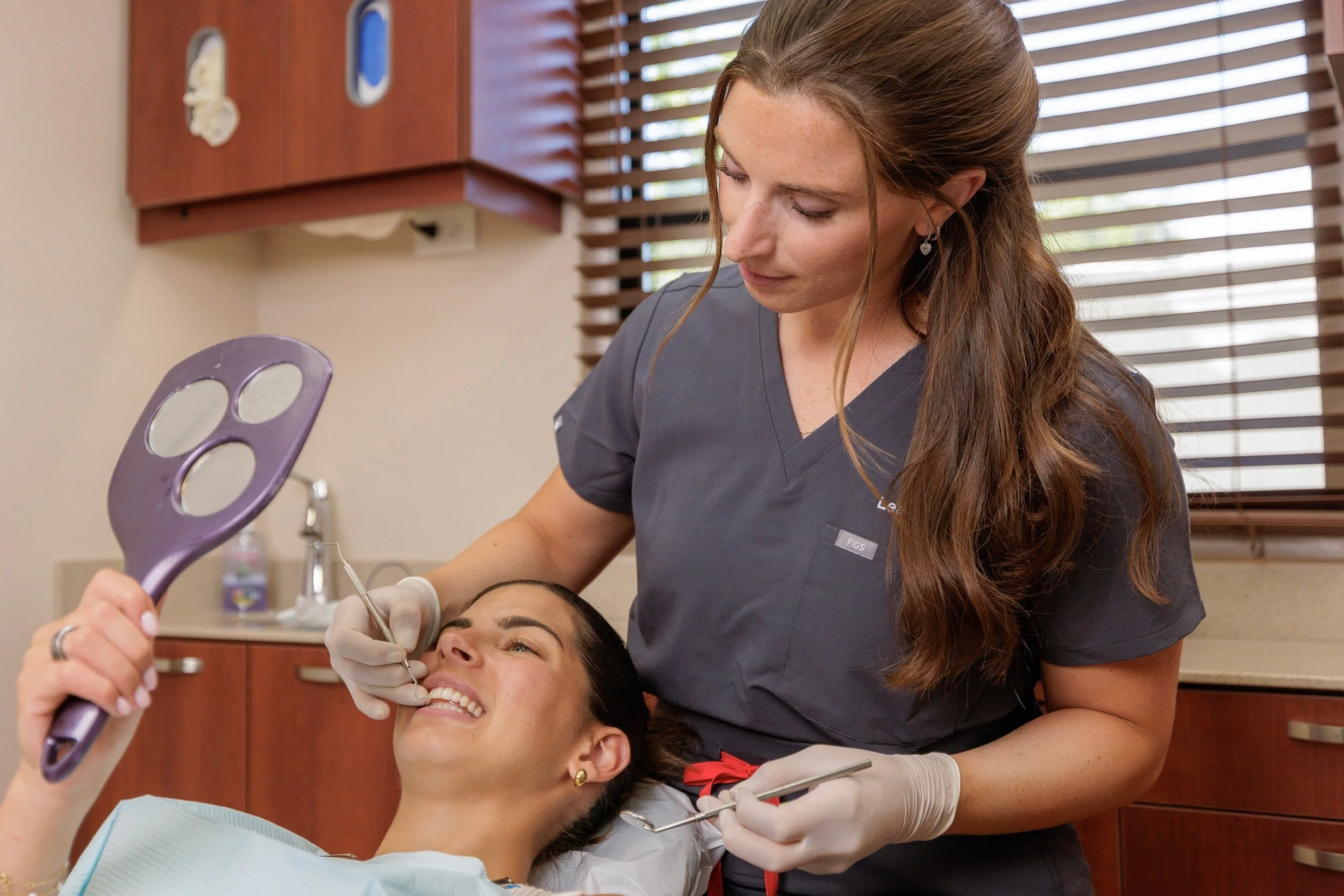A female dentist with long brown hair and earrings performs a dental procedure on a smiling female patient with dark hair, gold earrings, and a red bow on her shirt, in a dental office with wooden cabinets and window blinds.