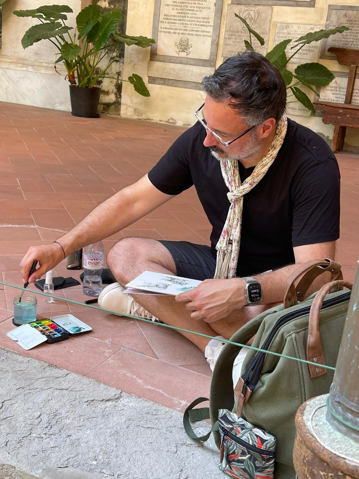 Un homme assis par terre avec un carnet, des pinceaux, une palette d'aquarelle, une bouteille d'eau et un flacon de désinfectant. Il semble peindre ou dessiner en extérieur, avec des plantes vertes en arrière-plan.