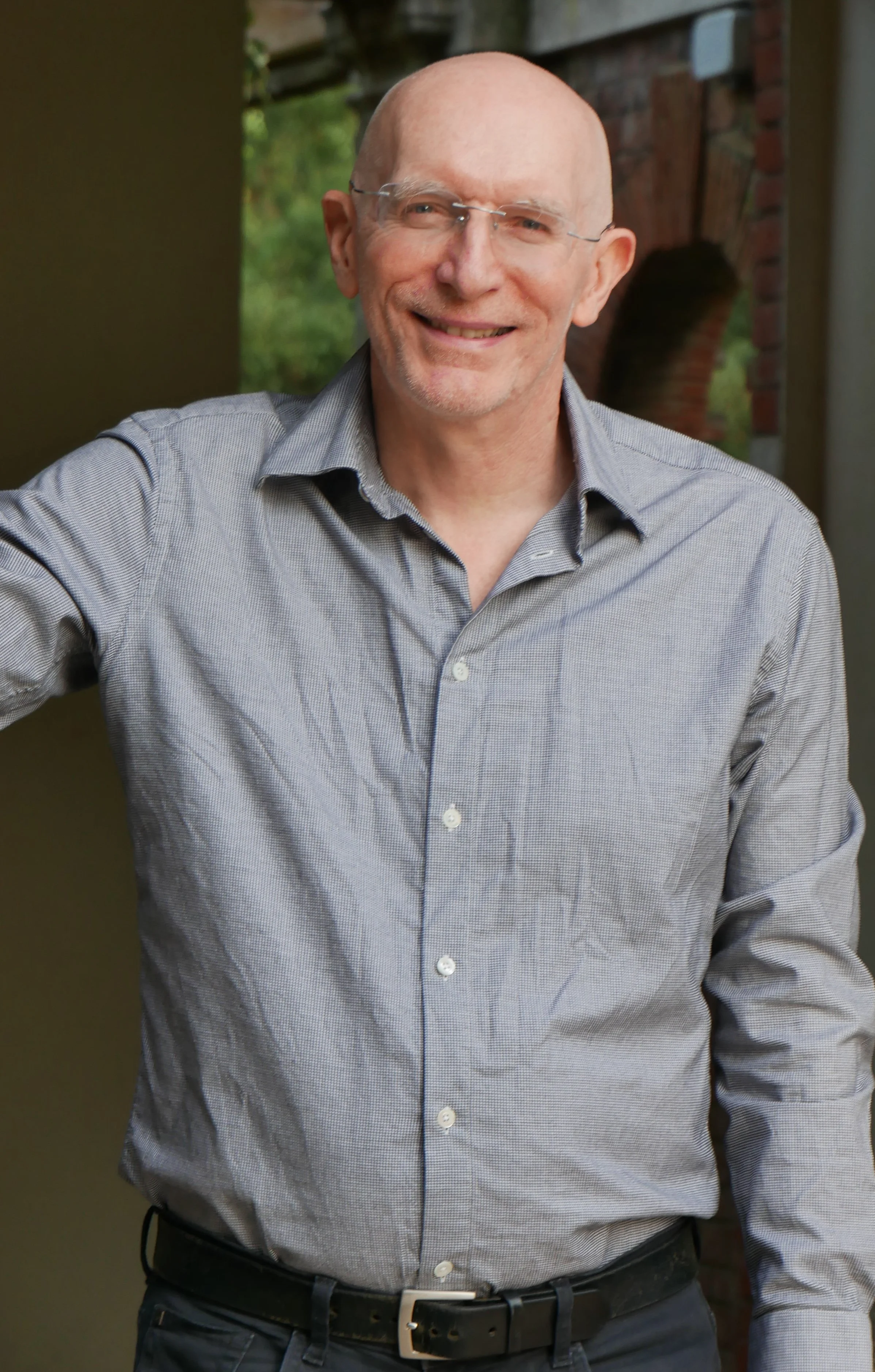 Richard Swynnerton, a smiling bald man wearing glasses and a grey shirt, standing outdoors with a leafy green background and a brick archway.