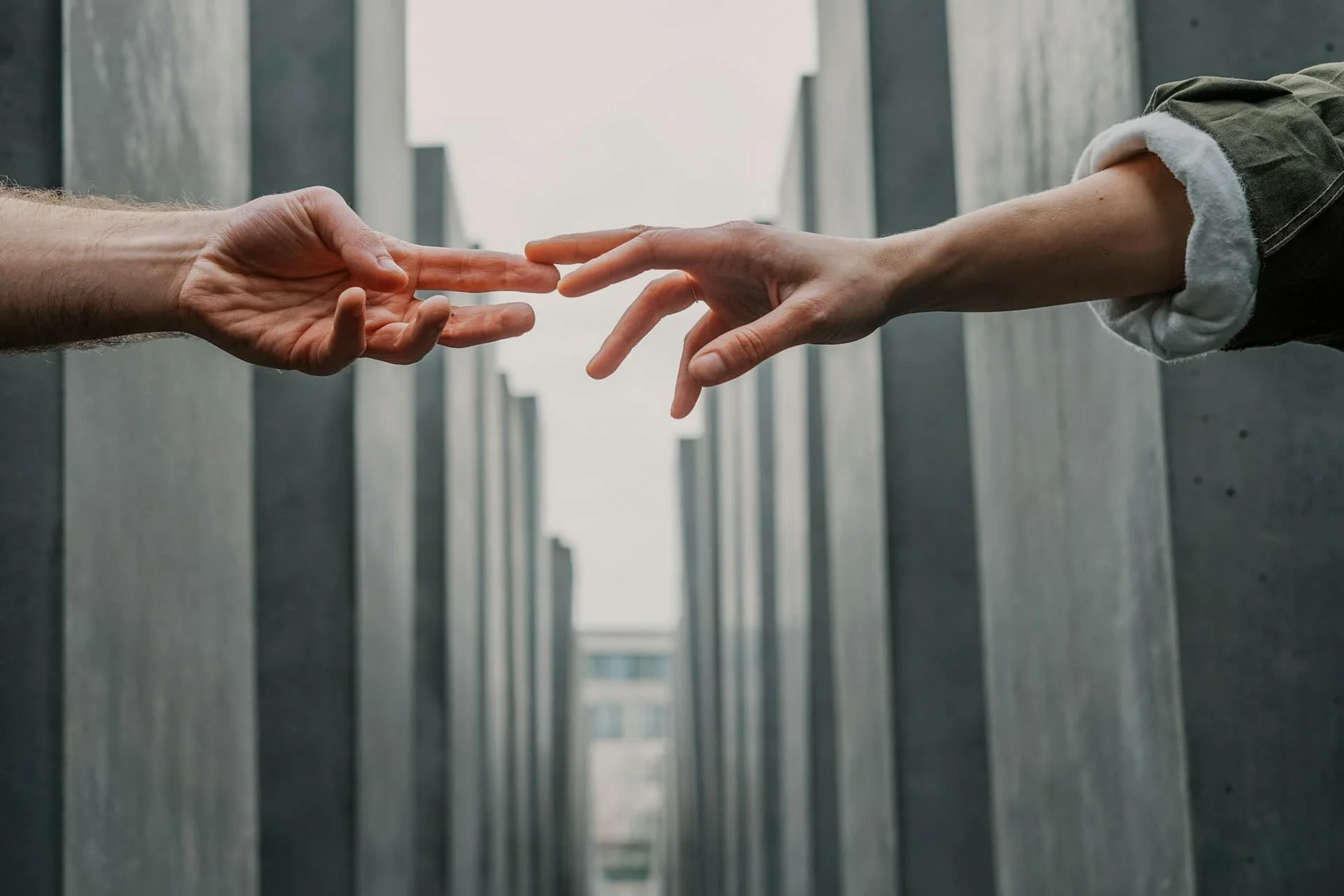 Two hands reaching towards each other with fingertips almost touching, framed between rows of tall concrete pillars, with a building visible in the distance.