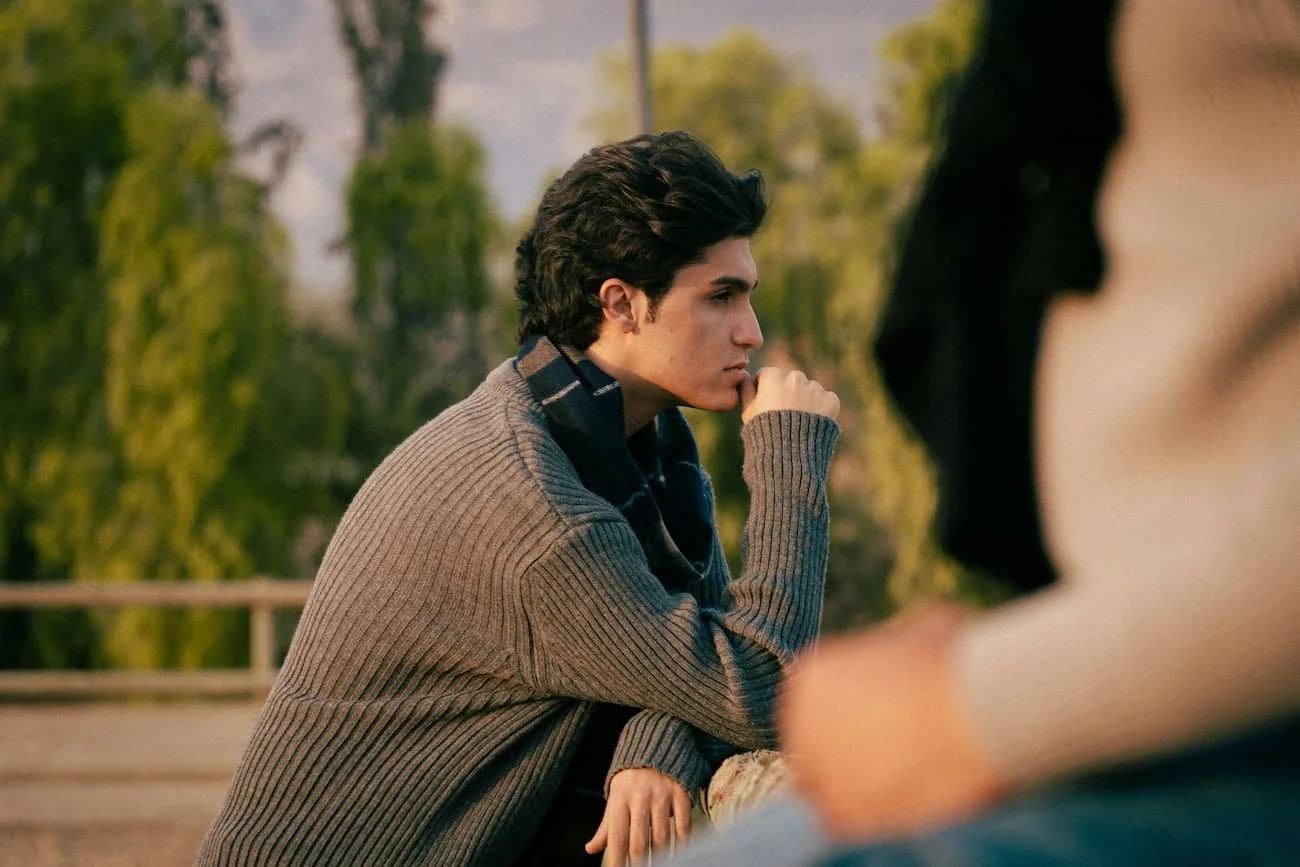 A young man in a grey ribbed sweater sitting outdoors, resting his chin on his hand with a thoughtful, withdrawn expression, trees in the background and another person partly visible beside him.