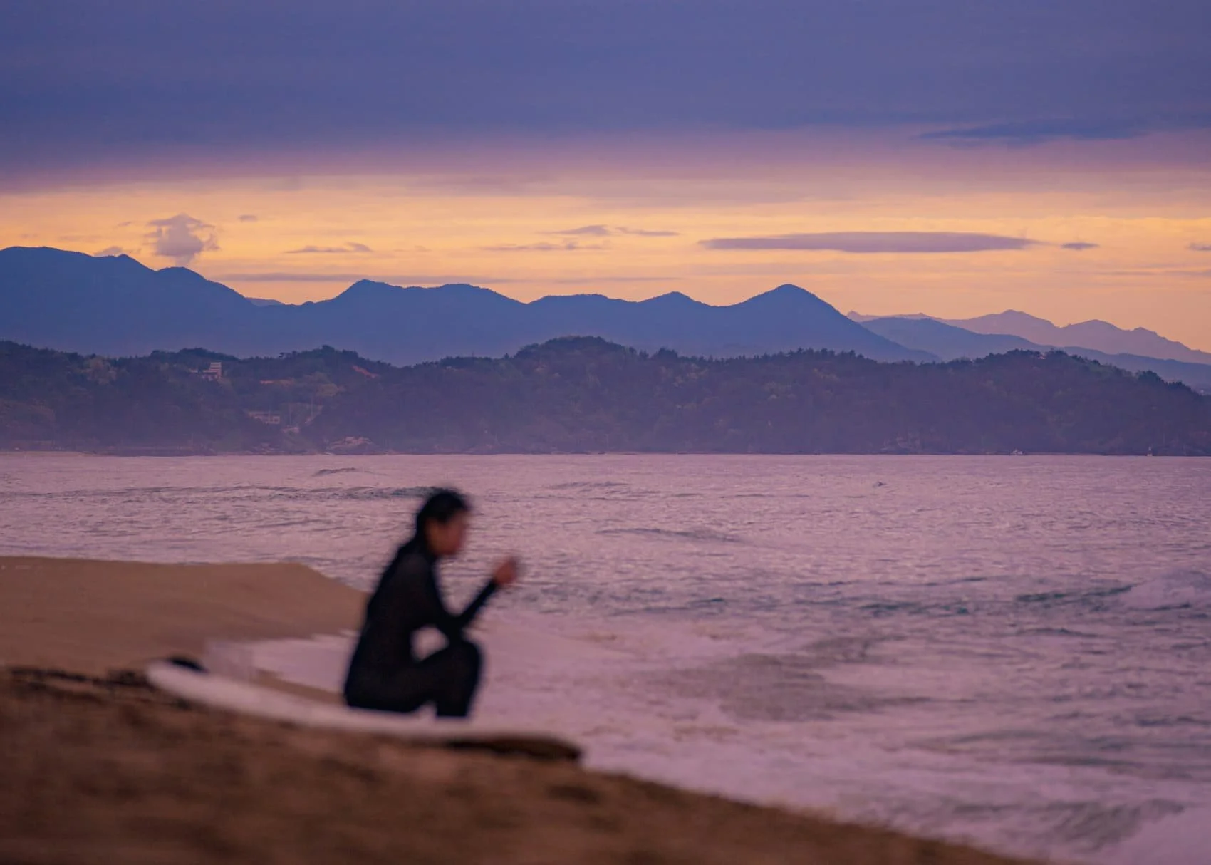 A person sitting alone on a beach at dusk, silhouetted against the sea with mountains and a purple-orange sky in the background.