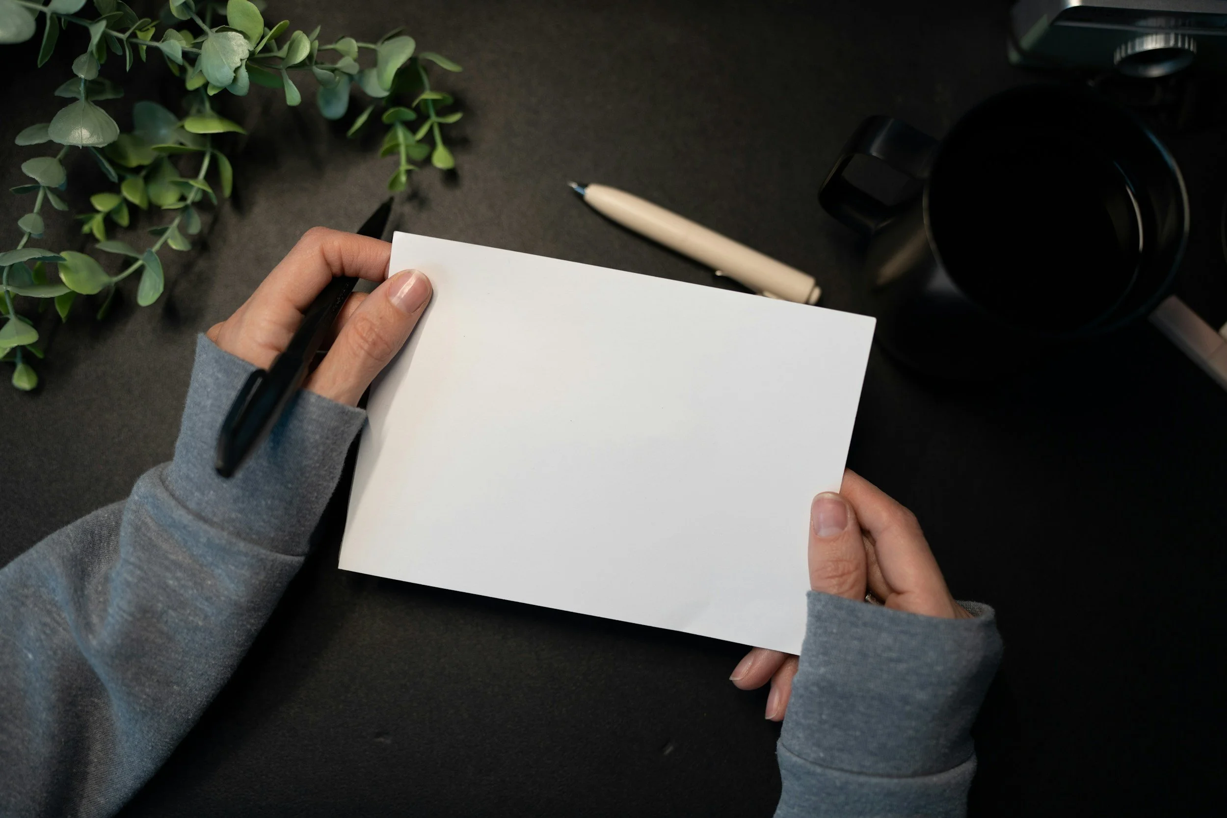 Person holding a blank white card with a black pen, surrounded by a green plant, a white pen, and black headphones on a dark surface.
