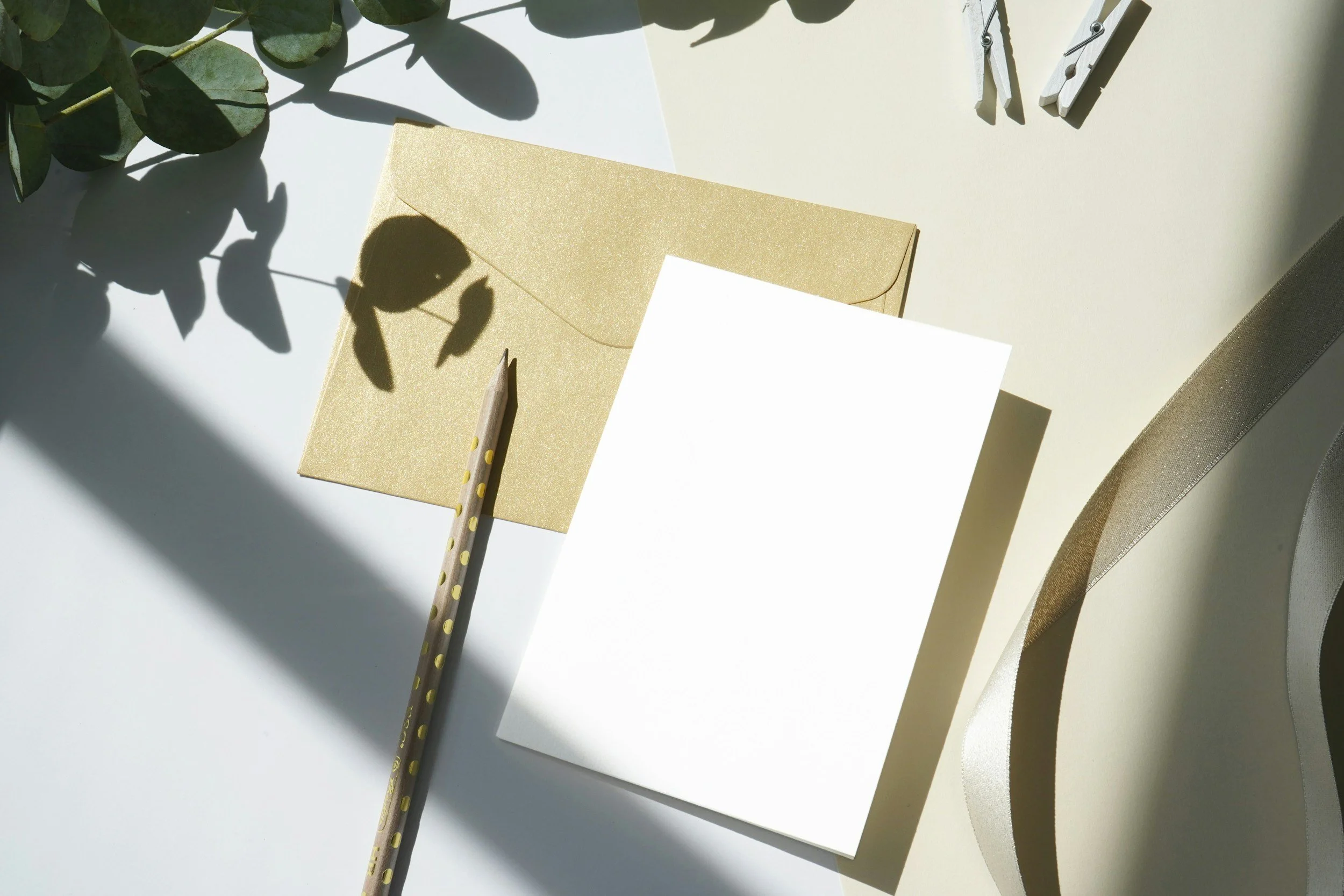 Flat lay of a workspace with a gold envelope, blank white card, yellow pencil with polka dots, green leafy branch, and a small wooden clothespin on a white surface, illuminated by sunlight and shadows.