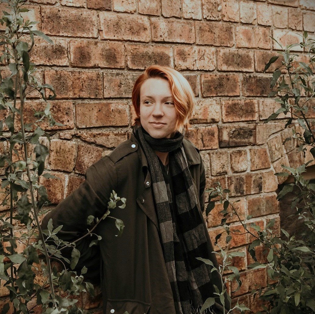 A woman with short red hair leaning against a brick wall surrounded by green foliage, wearing a brown jacket and a black and gray checkered scarf.
