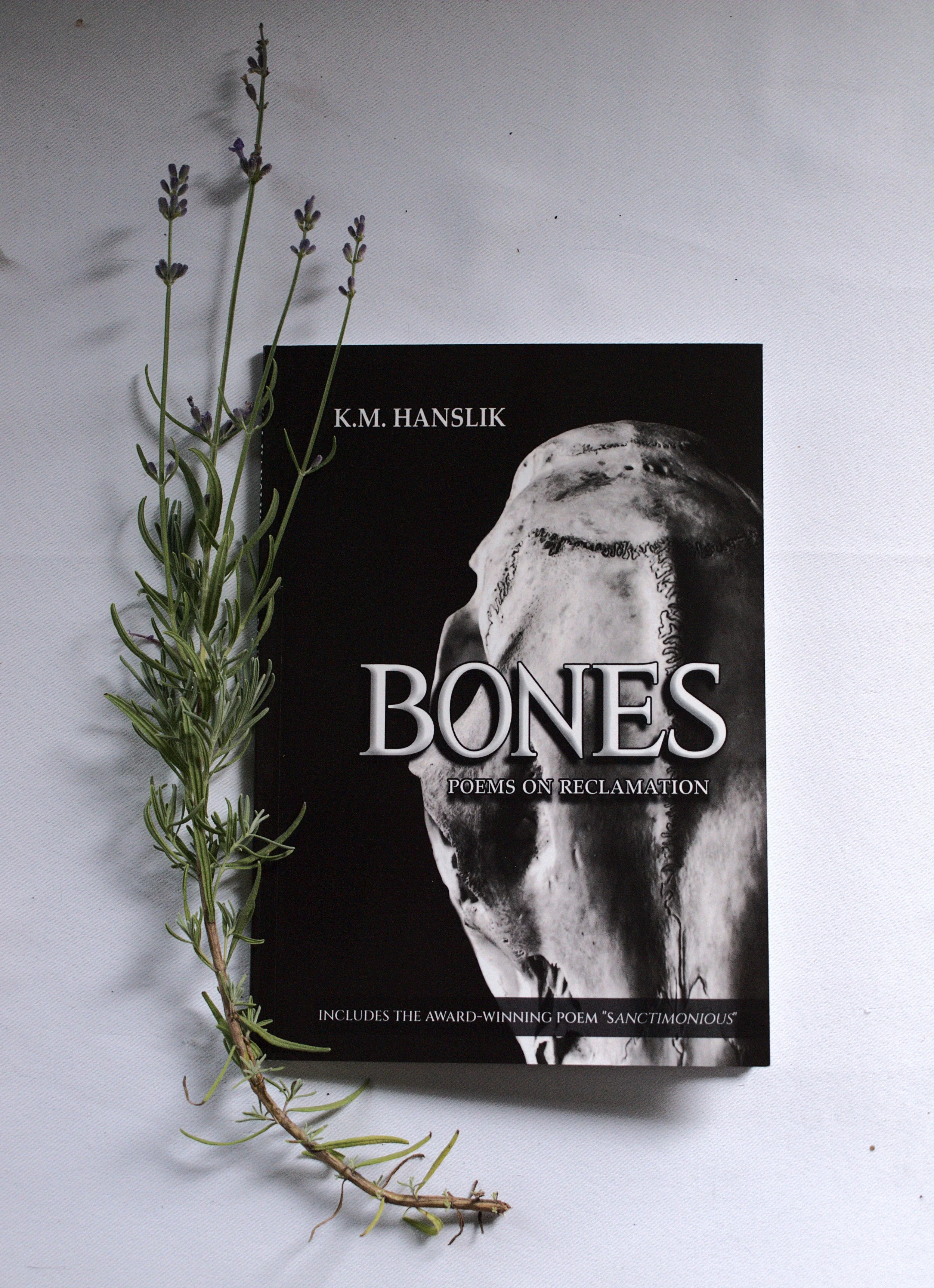 A book titled 'Bones' by K.M. Hanslik, with a black and white skull illustration on the cover, is placed on a white surface. A sprig of purple-flowered herb, likely lavender, is laid diagonally across the top left corner of the book.